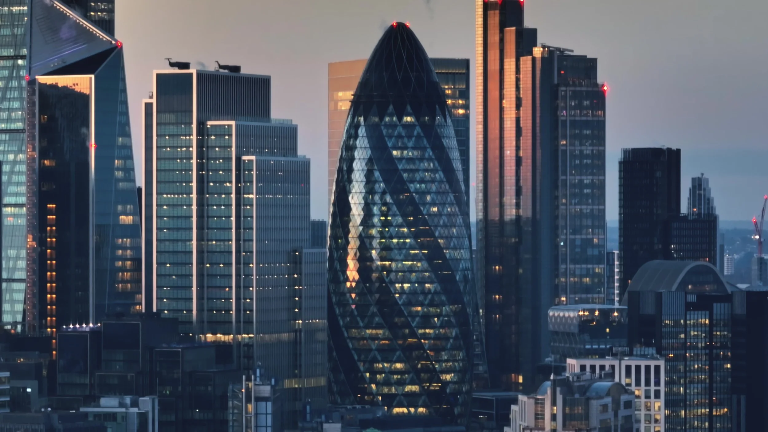 Skyline of London with prominent modern skyscrapers, including the Gherkin and the 'walkie-talkie' building, during evening with city lights illuminated.