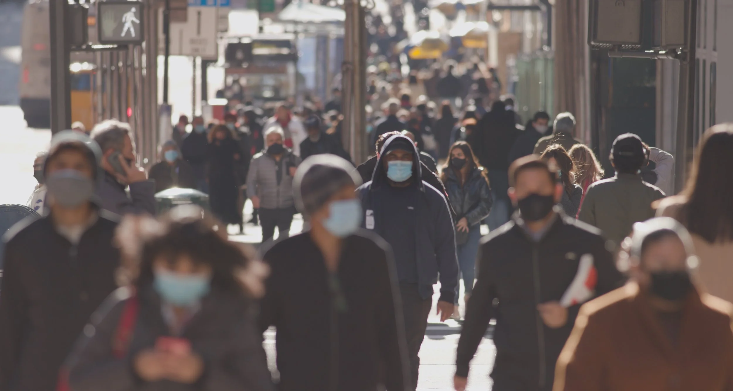 A crowded city street with pedestrians wearing masks walking in various directions.