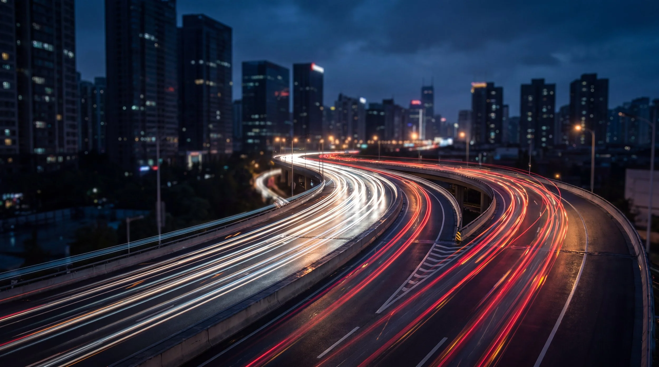 Nighttime cityscape with light trails from moving vehicles on a curved highway, surrounded by tall dark buildings.