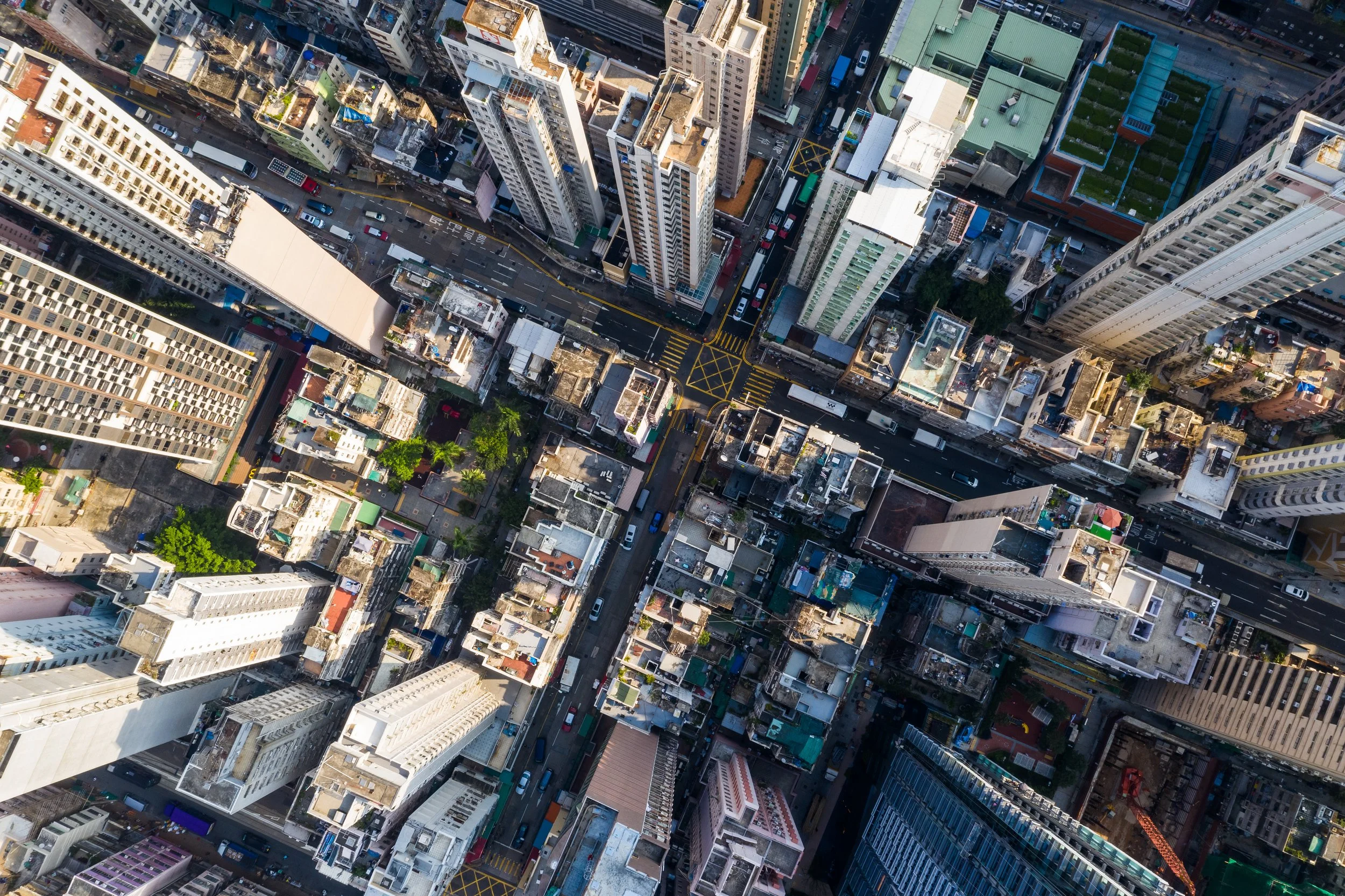 An aerial view of a densely packed city with high-rise buildings, streets, and parked cars. The buildings vary in height and design, and the streets are marked with yellow and white lines.