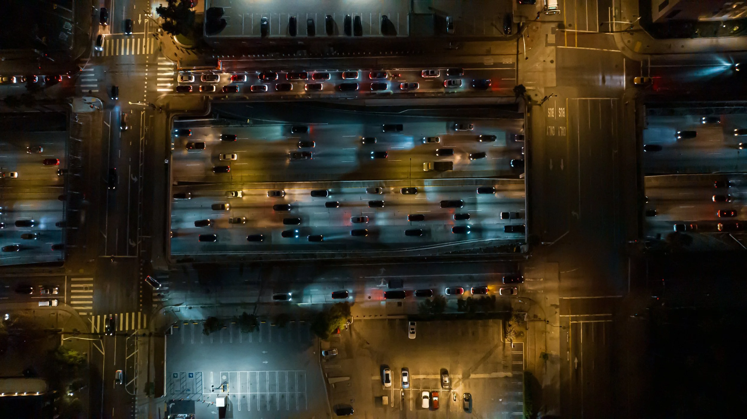 An aerial view of a city street at night with multiple lanes of traffic, illuminated by streetlights and vehicle headlights. There are parked cars, crosswalks, and traffic signals visible, with some cars moving and others stopped.