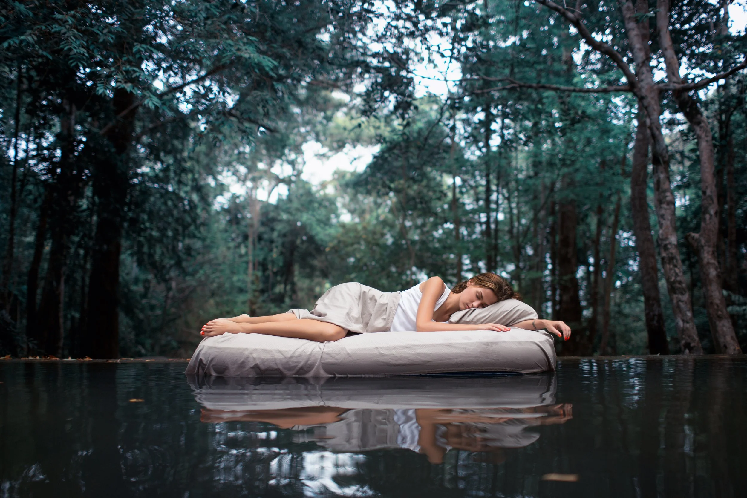A woman sleeping on a bed placed on a body of water in a forest setting.