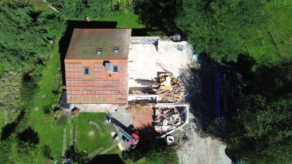 Luftaufnahme eines Abbruchgrundstücks mit einem teildemontierten Wohnhaus, freigelegtem Erdgeschossbereich, Bauschutthalden und einem Mobilbagger.