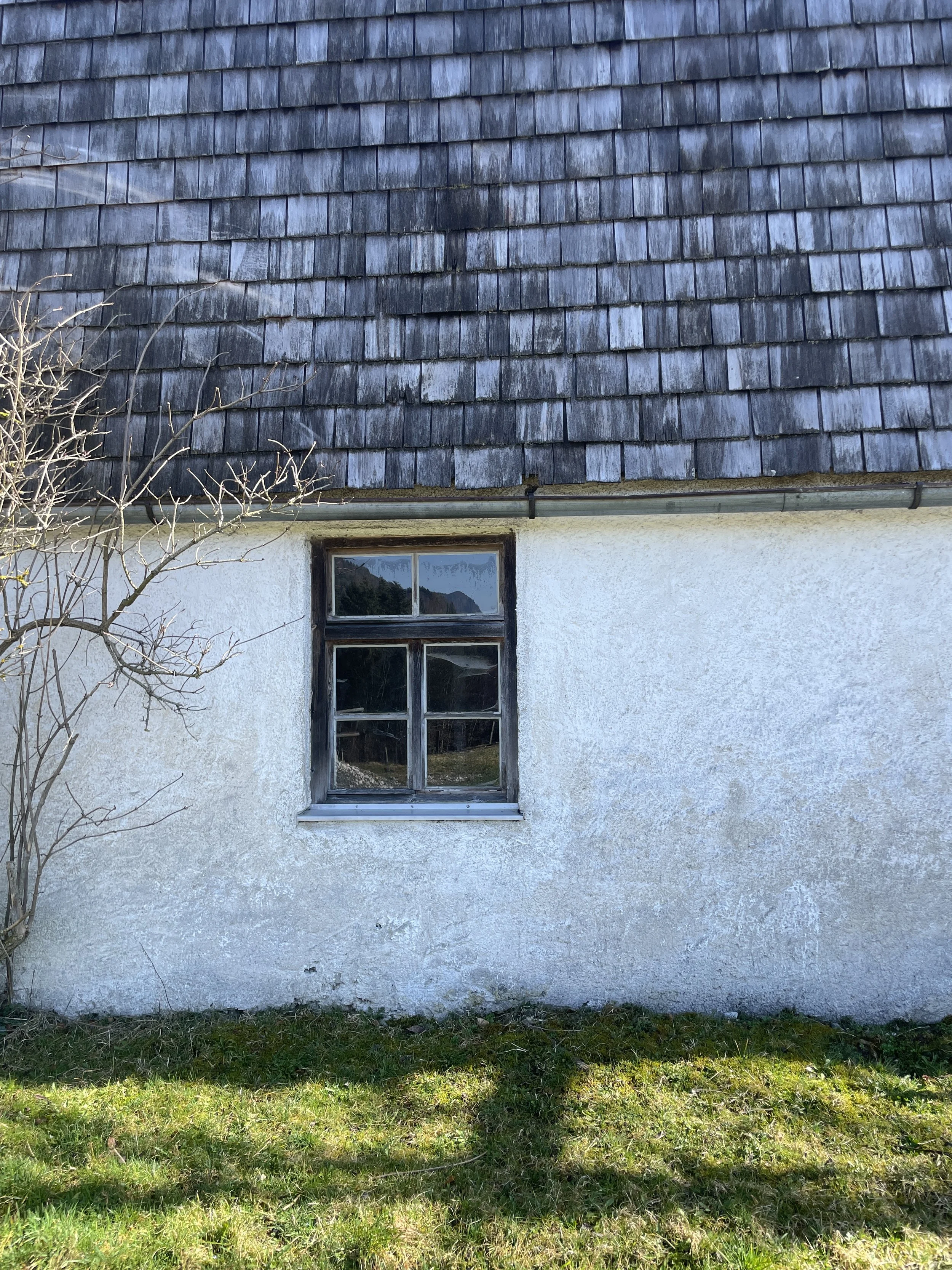 Detailaufnahme einer weißen Putzfassade eines historischen Einfirsthofs mit dunklem Sprossen-Holzfenster und verwitterten Holzschindeln am Dachansatz.