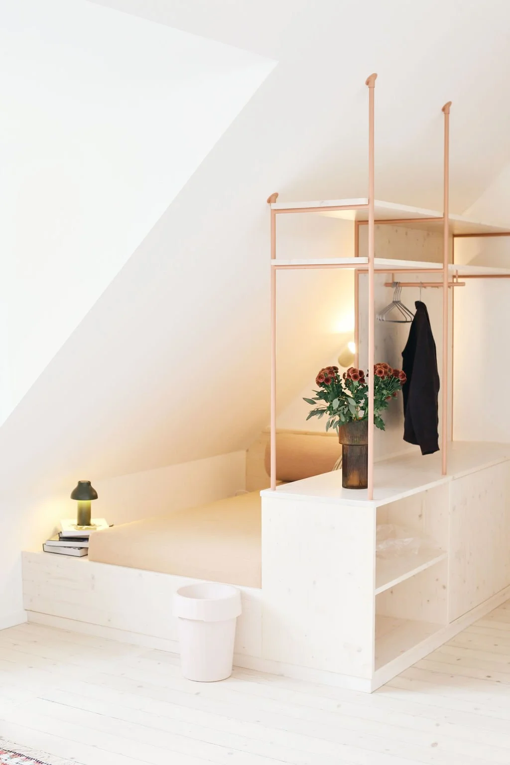 Maßgefertigter Einbau unter einer Dachschräge mit integriertem Podestbett, rosafarbenem filigranem Regalgestell als Raumteiler, Garderobenstange und stilvoller Beleuchtung.