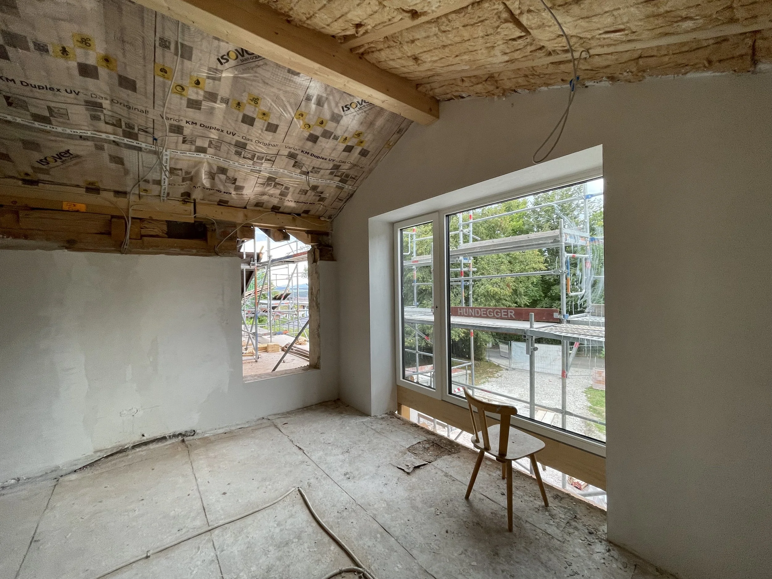 Dachgeschoss-Baustelle mit freigelegtem Holzgebälk, sichtbarer Zwischensparrendämmung mit Dampfbremsfolie, frisch verputzter Wand und bodentiefem Fenster.