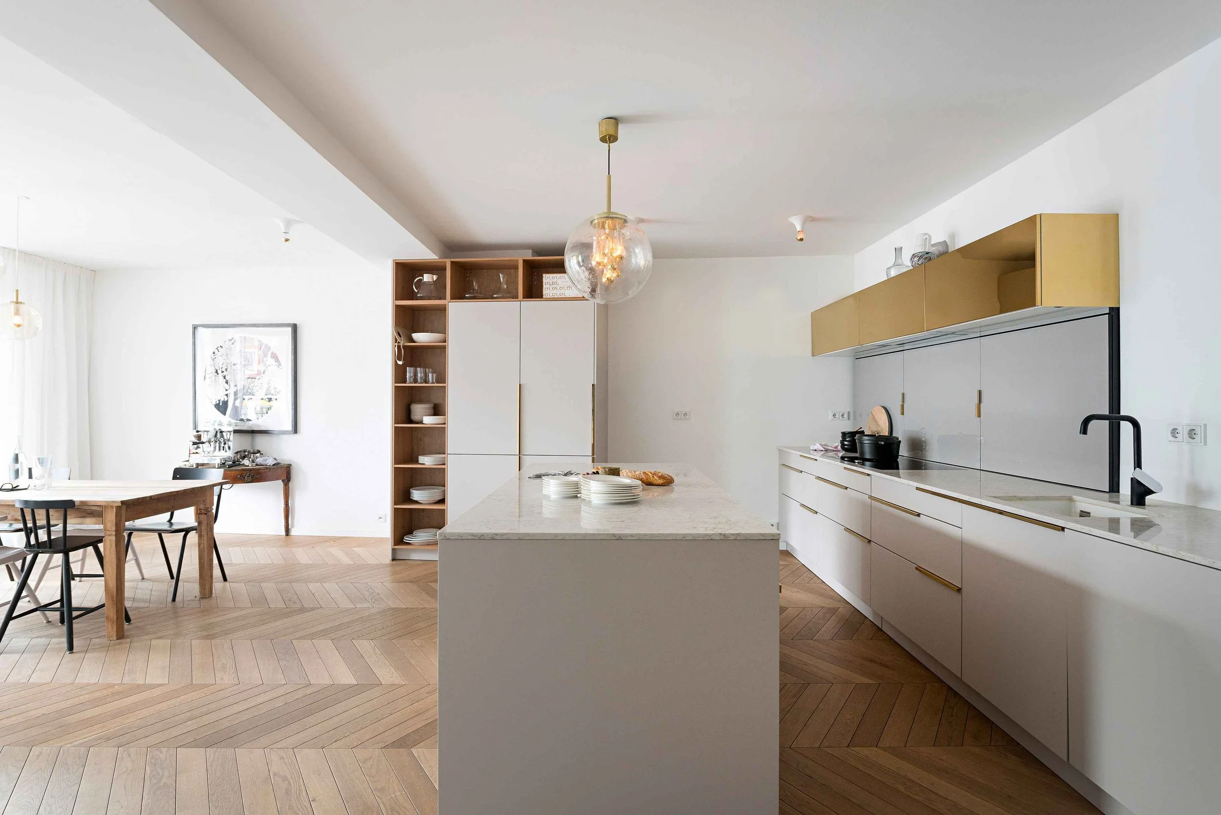 Moderne, helle Küche mit weißer Kücheninsel, Marmorarbeitsplatte, messingfarbenen Hängeschränken und Fischgrät-Parkett im Sanierungsprojekt in München.