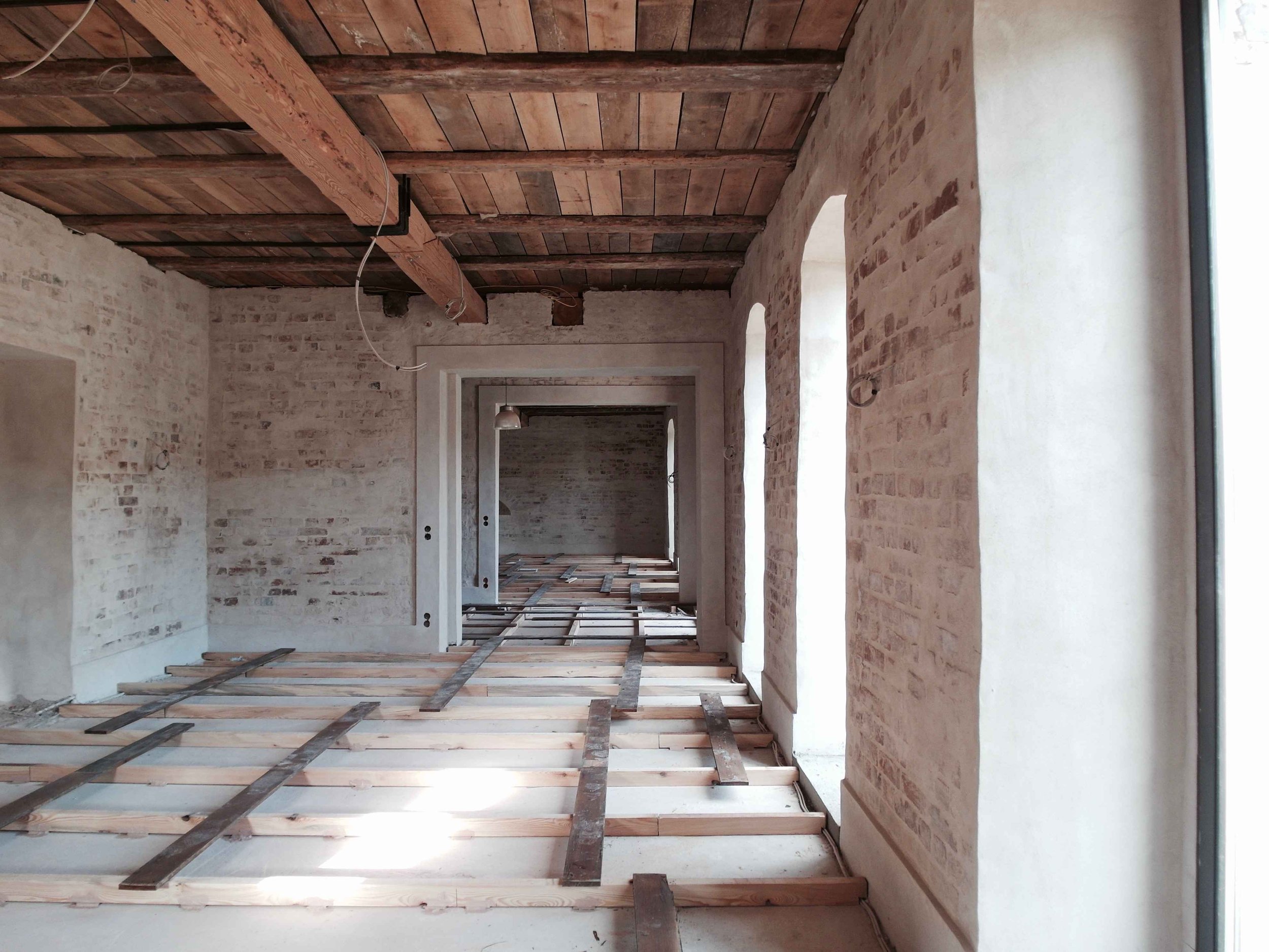 Innenansicht eines langen, im Bau befindlichen Raumes mit offen liegenden Holzdeckenbalken, unverputzten Backsteinwänden und einer provisorischen Unterkonstruktion für den Fußboden aus Holzbalken auf Beton.