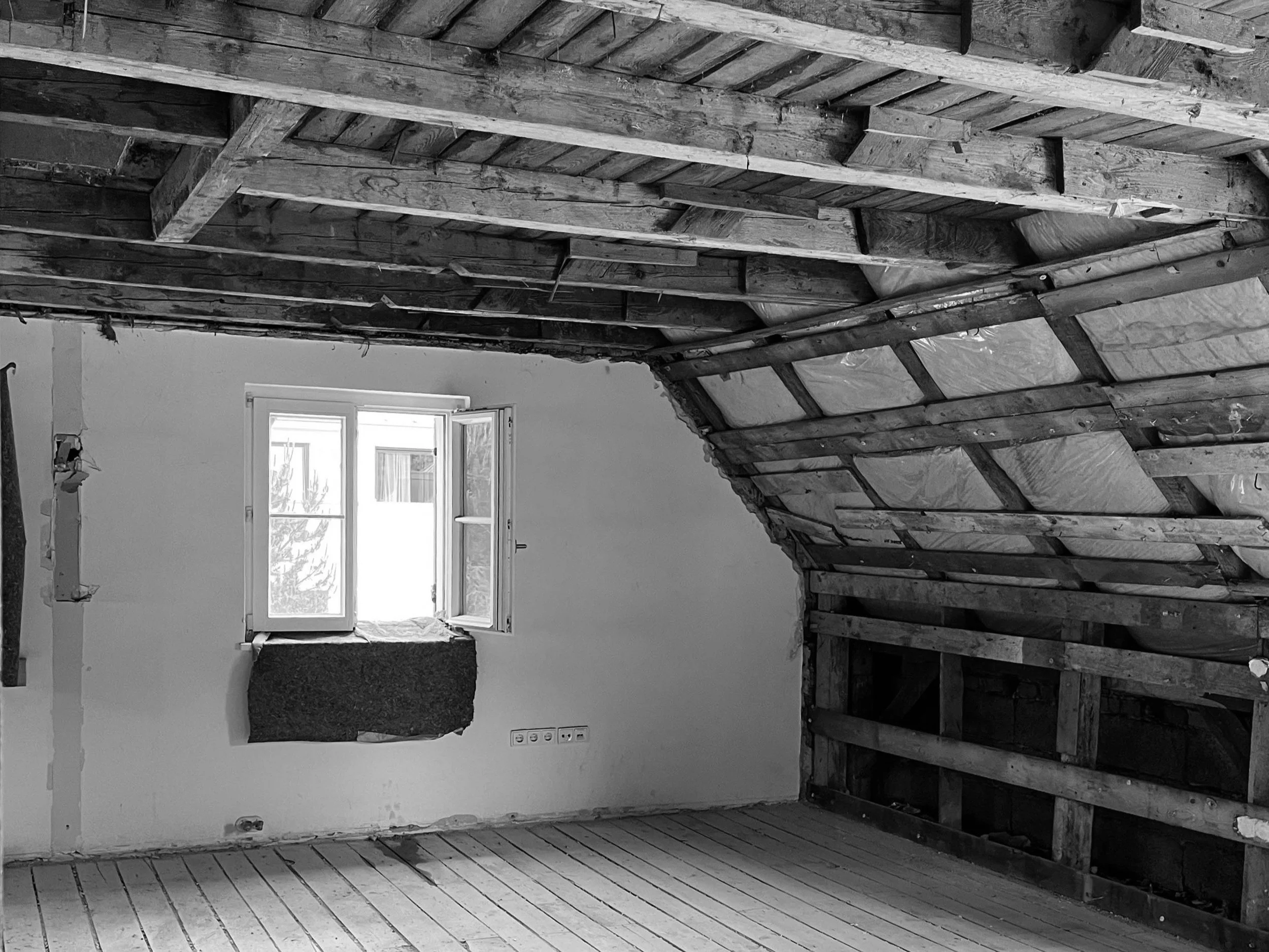 Aufnahme eines Dachraums im Rohbau-Zustand während der Sanierung mit freigelegter Holz-Dachkonstruktion, geöffneter Dachschräge, einfachem Dielenboden und geöffnetem Fenster.