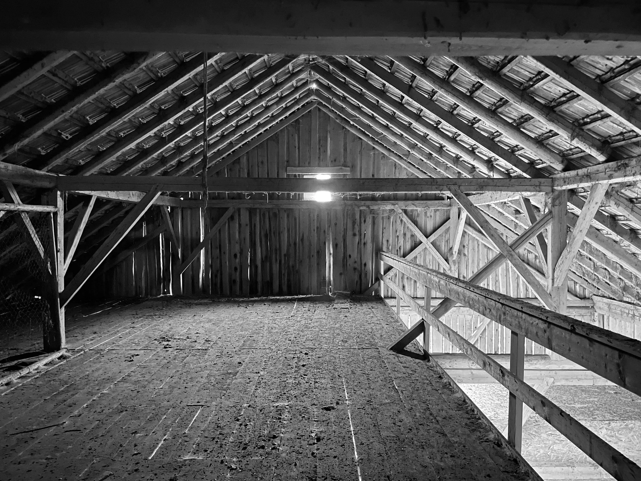 Aufnahme eines riesigen, unrenovierten Scheunendachbodens mit freiliegender Holz-Dachkonstruktion, sichtbaren Dachlatten, Holzgeländer und Lichteinfall an der Stirnseite.
