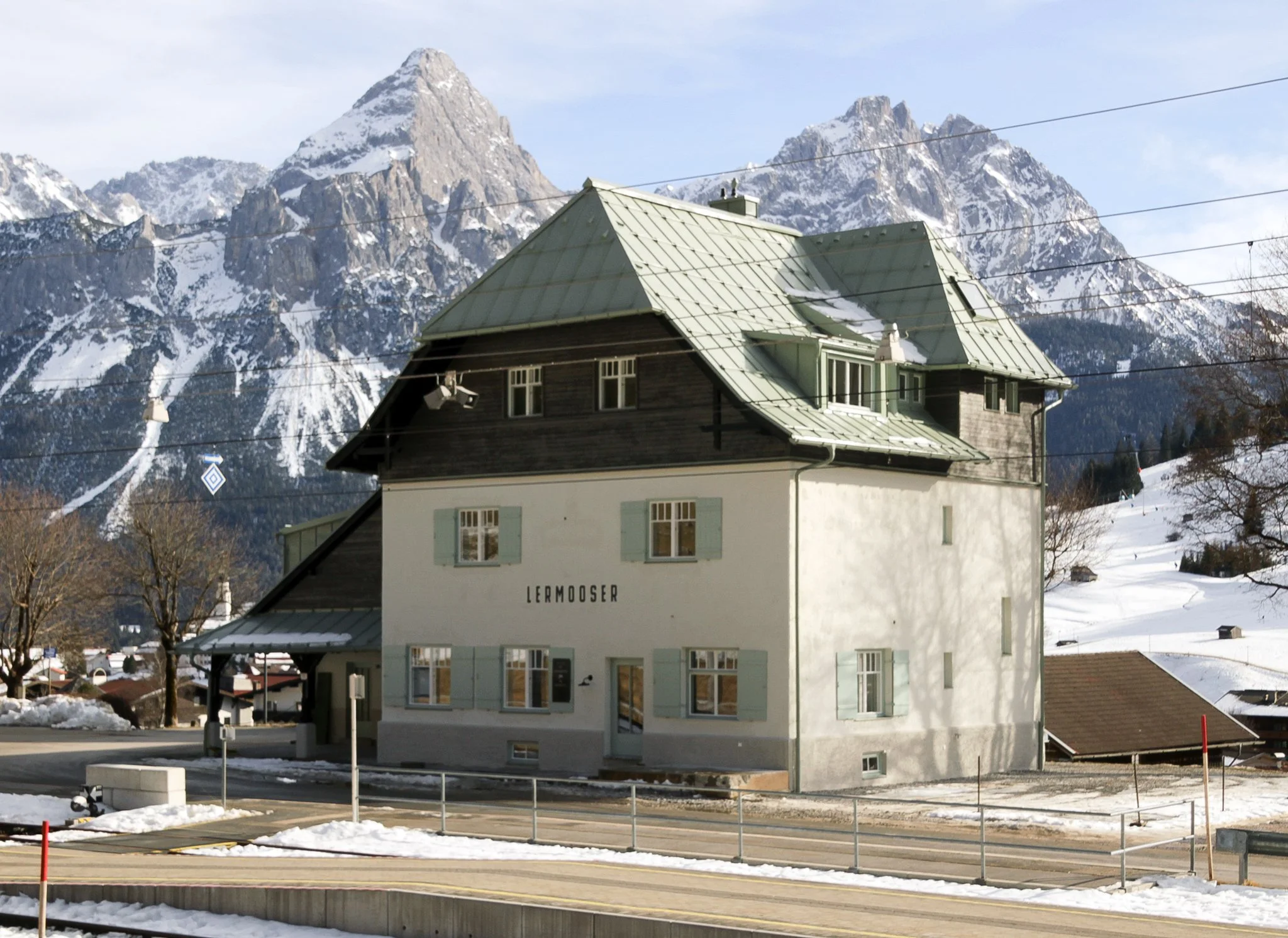 Außenansicht des historischen, denkmalgeschützten Bahnhofsgebäudes Lermoos mit grünem Blechdach, Holzschindeln im Dachgeschoss und hellgrünen Klappläden vor verschneiter Bergkulisse der Zugspitzregion.