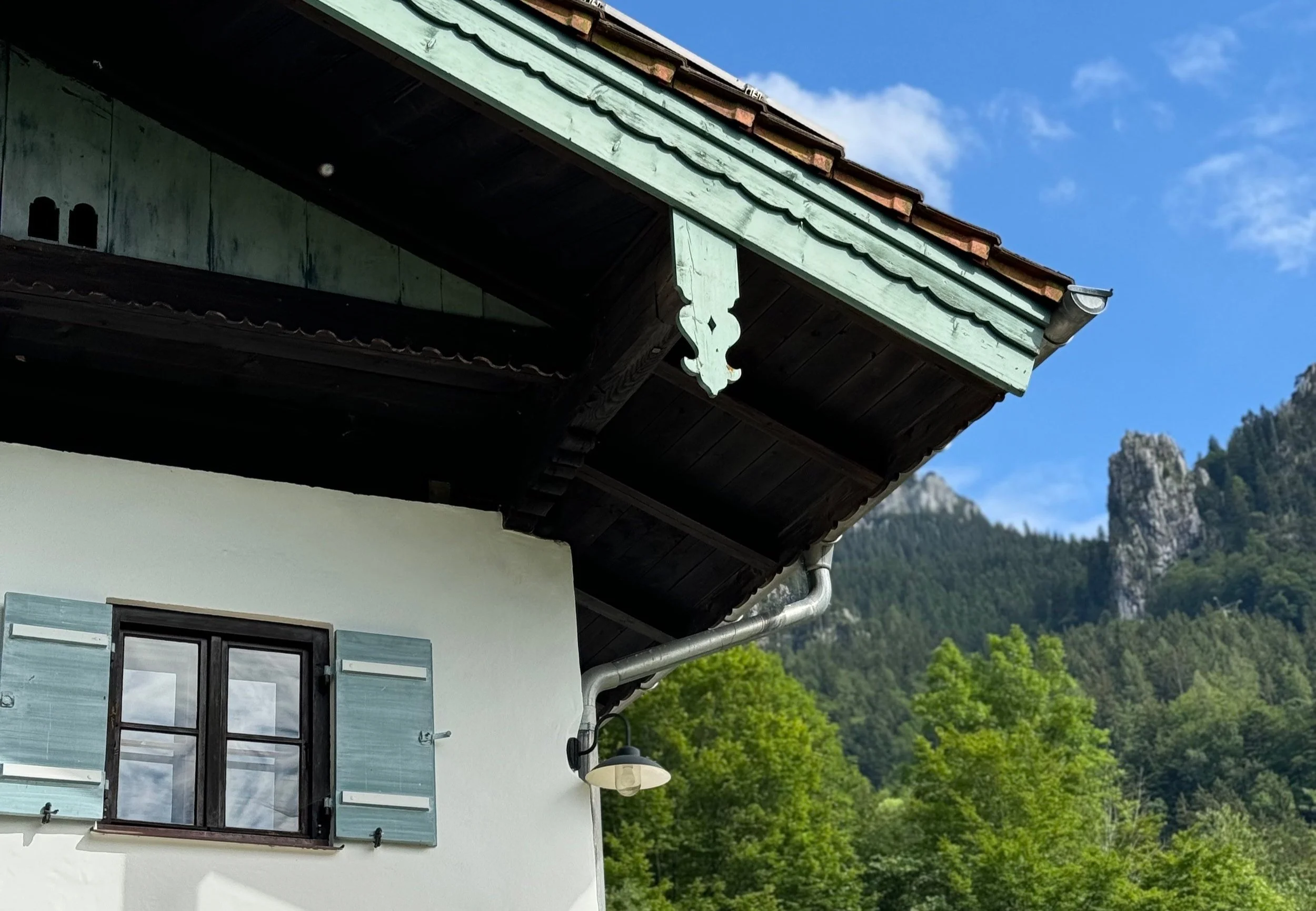 Detailaufnahme eines traditionellen alpenländischen Bauernhauses mit hellgrün verzierter Windfeder am Dachüberstand, hellblauen Holz-Klappläden und Sprossenfenster vor Bergkulisse.