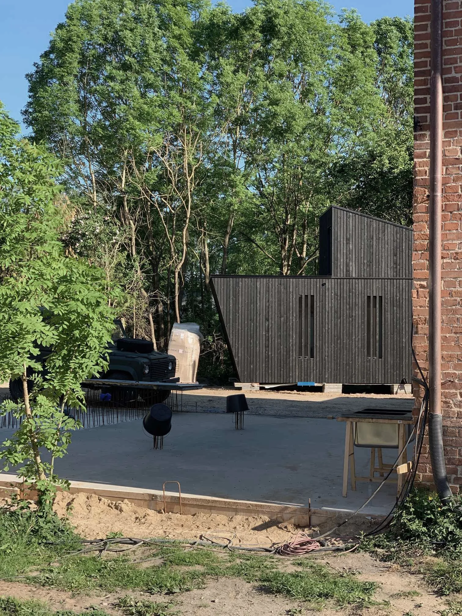 Außenaufnahme einer Baustelle mit einem aufgestellten, modernen Modulhaus aus dunklem Holz, Fundamentplatte im Vordergrund und Bäumen im Hintergrund.