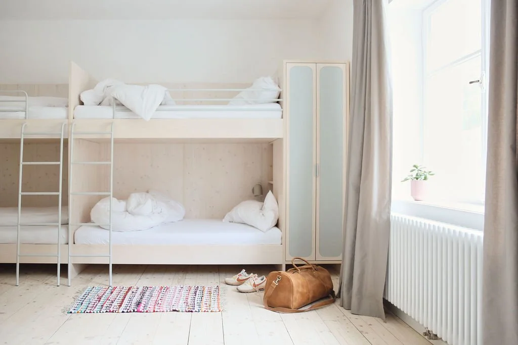 Hell eingerichtetes Hotelzimmer mit eingebauten Doppel-Etagenbetten aus hellem Holz, Holzdielenboden, Schrank und großem Fenster mit hellen Vorhängen.