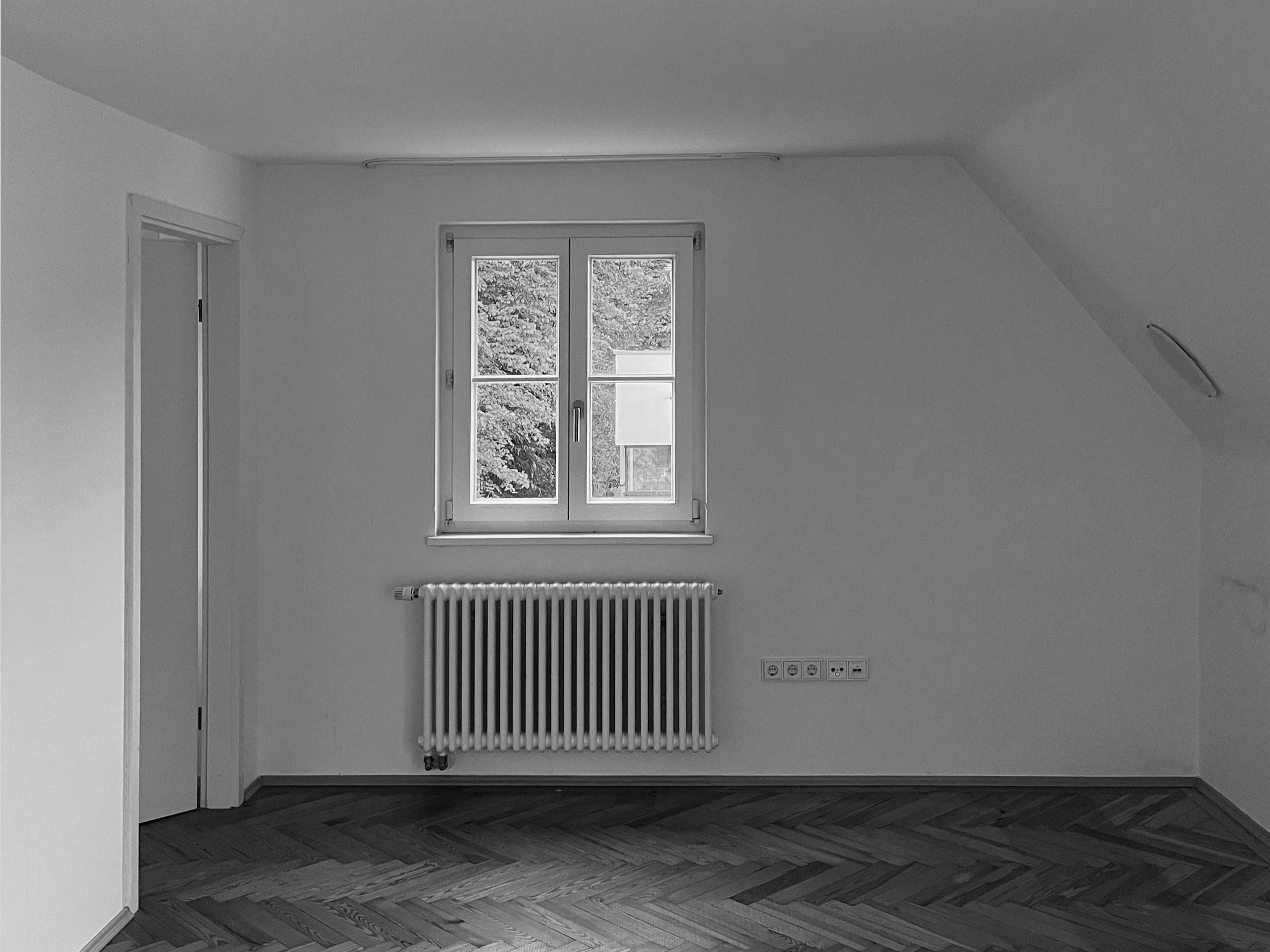 Aufnahme eines Dachgeschoss-Raums vor der Sanierung mit weiß gestrichenen Wänden, Fischgrät-Parkett, einem zweiflügeligen Fenster über dem Heizkörper und einer geöffneten Innentür.
