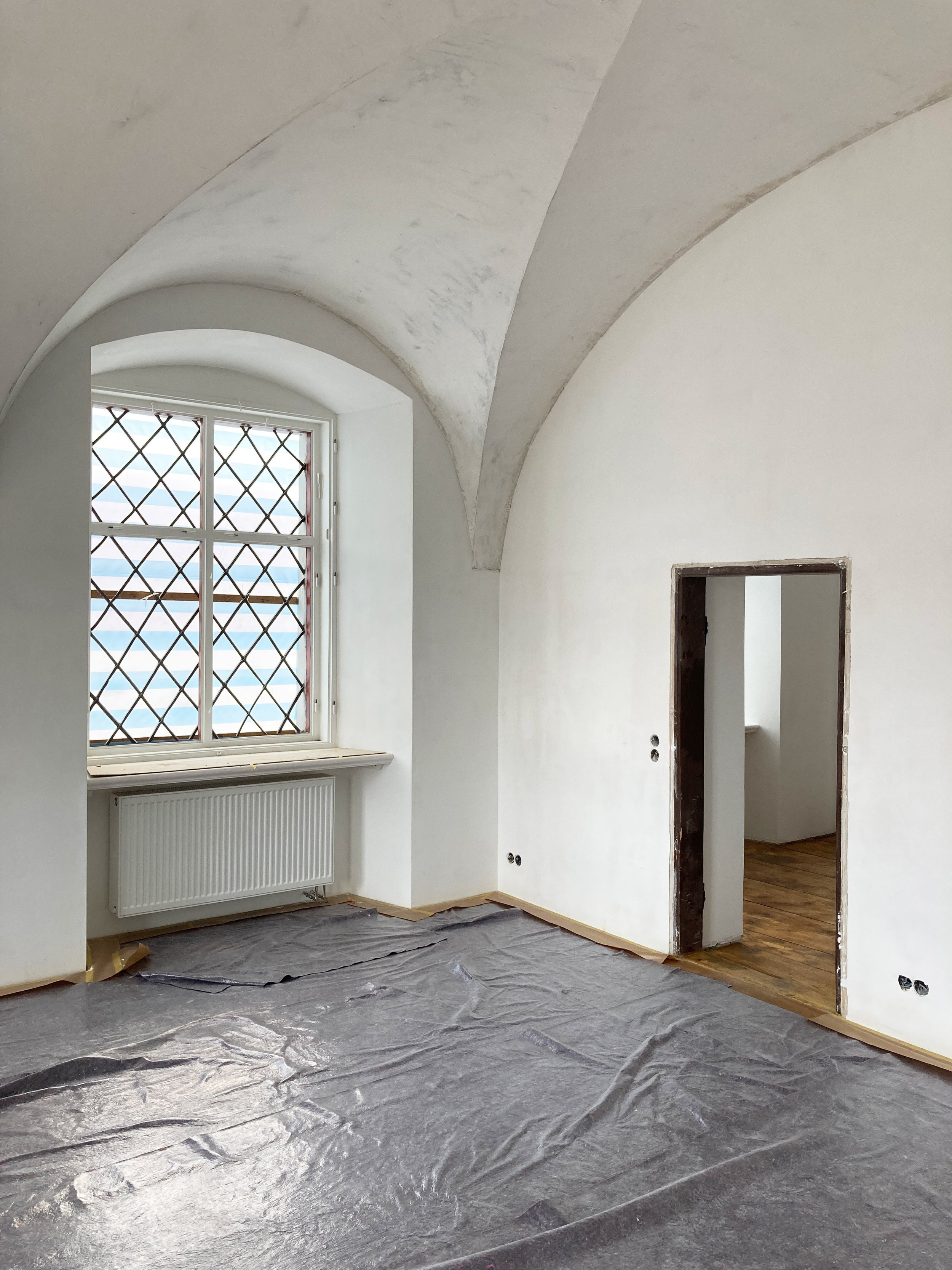 Frisch verputzter Schlossraum während der Renovierung mit hellem Gewölbe, rautenförmig geteiltem Bleiglasfenster und ausgelegter Schutzfolie am Boden.