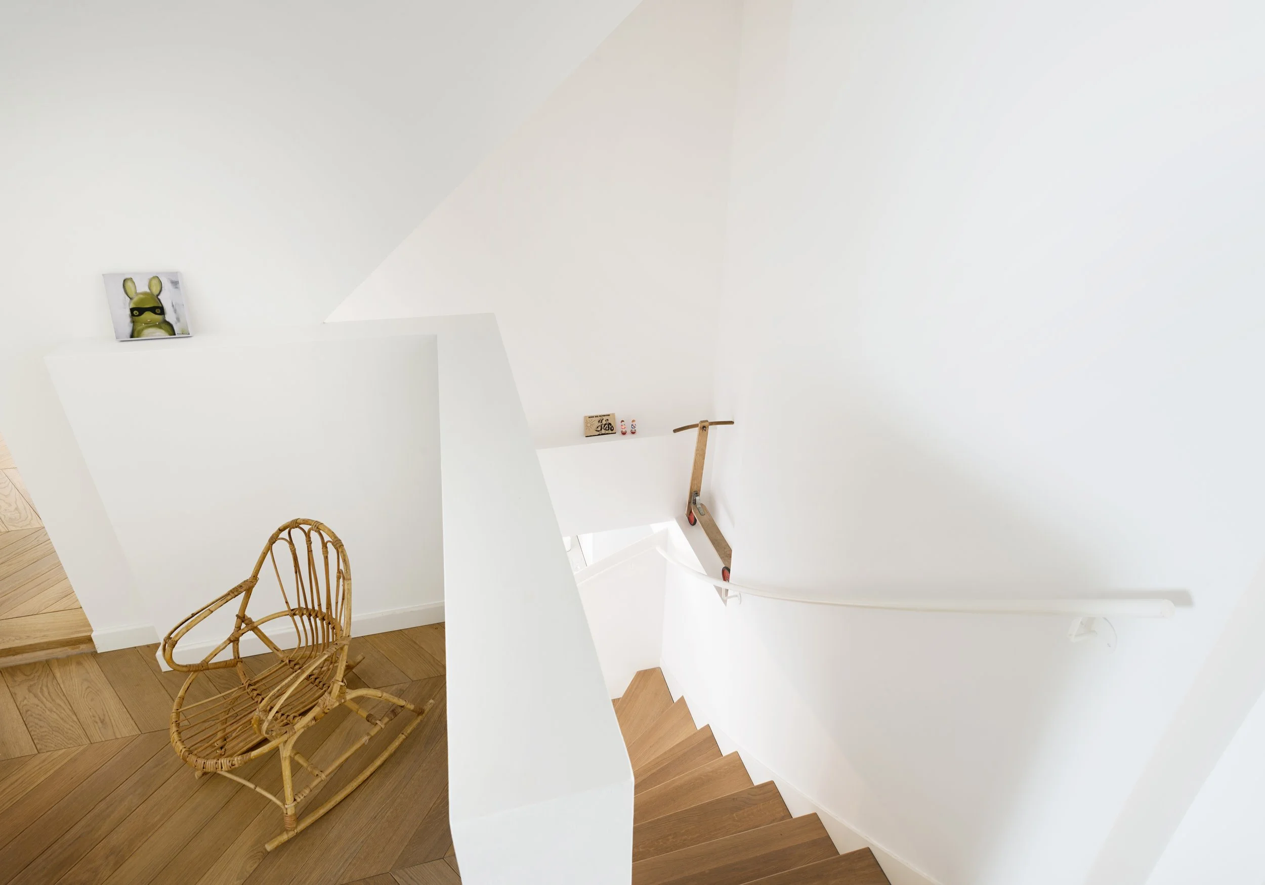 Blick von oben auf eine weiße Treppe mit Holzstufen, Fischgrät-Parkett auf der Galerie und einem kleinen Rattan-Schaukelstuhl.