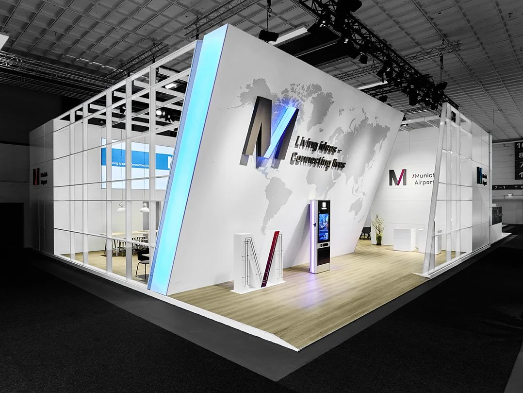 Außenansicht des freistehenden, modernen Messestands des Flughafens München mit beleuchteter Markenwand, Logo, Weltkartengrafik und Holzboden im Messehallen-Ambiente.