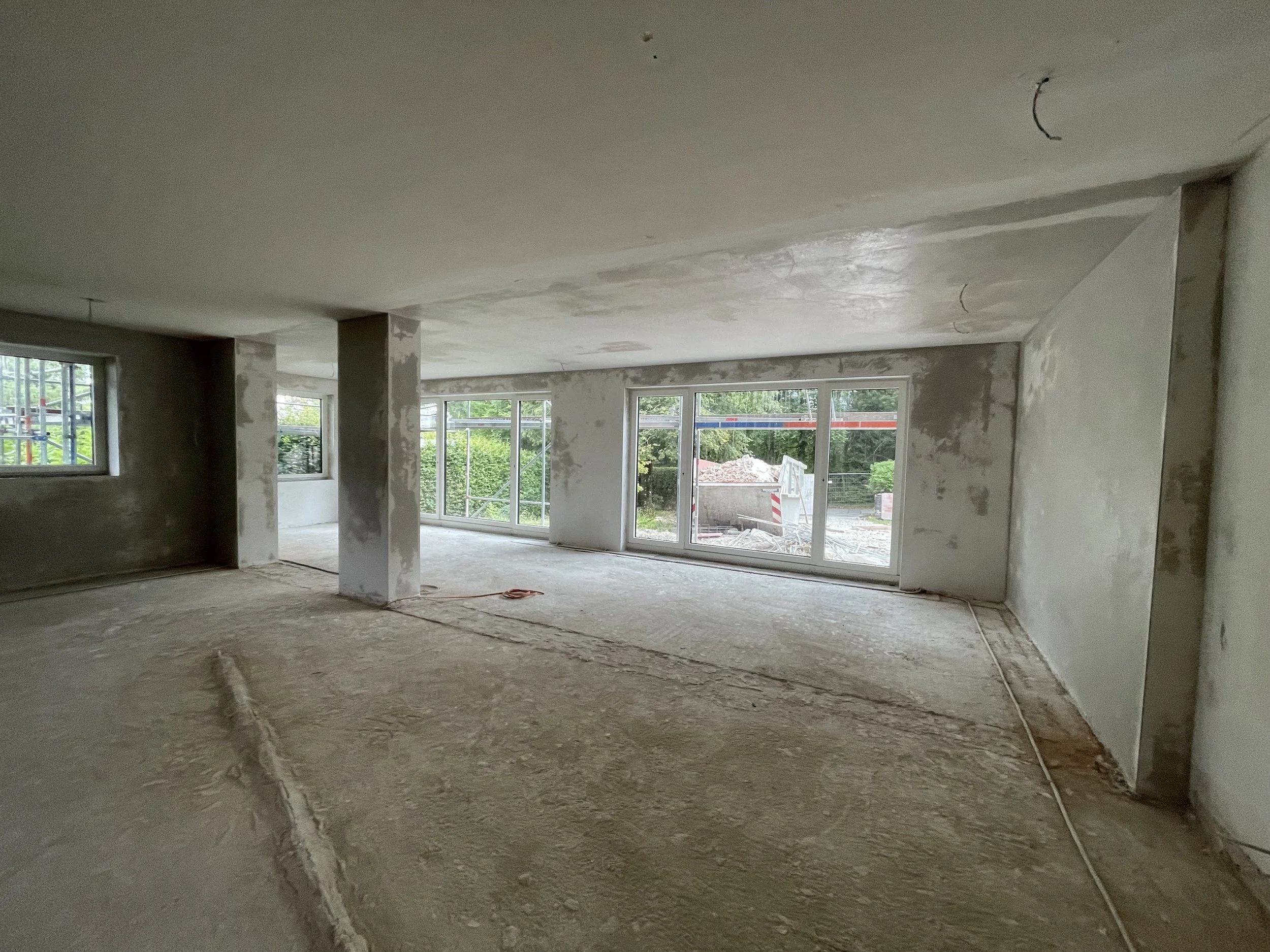 Baustellenfoto eines großzügigen Wohnraums in der Rohbauphase mit verputzten Innenwänden, Betonstütze, Rohfußboden und großen Schiebetürelementen zum Garten.