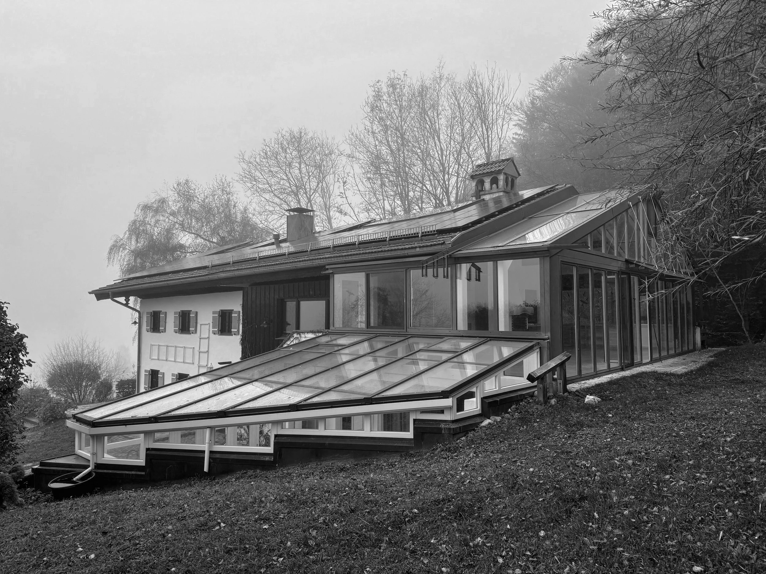 Aufnahme vor der Sanierung/dem Umbau eines Wohnhauses in Hanglage mit einem großflächigen, kaskadenförmigen Glasanbau (Wintergarten/Gewächshaus) und Photovoltaikanlage auf dem Dach.