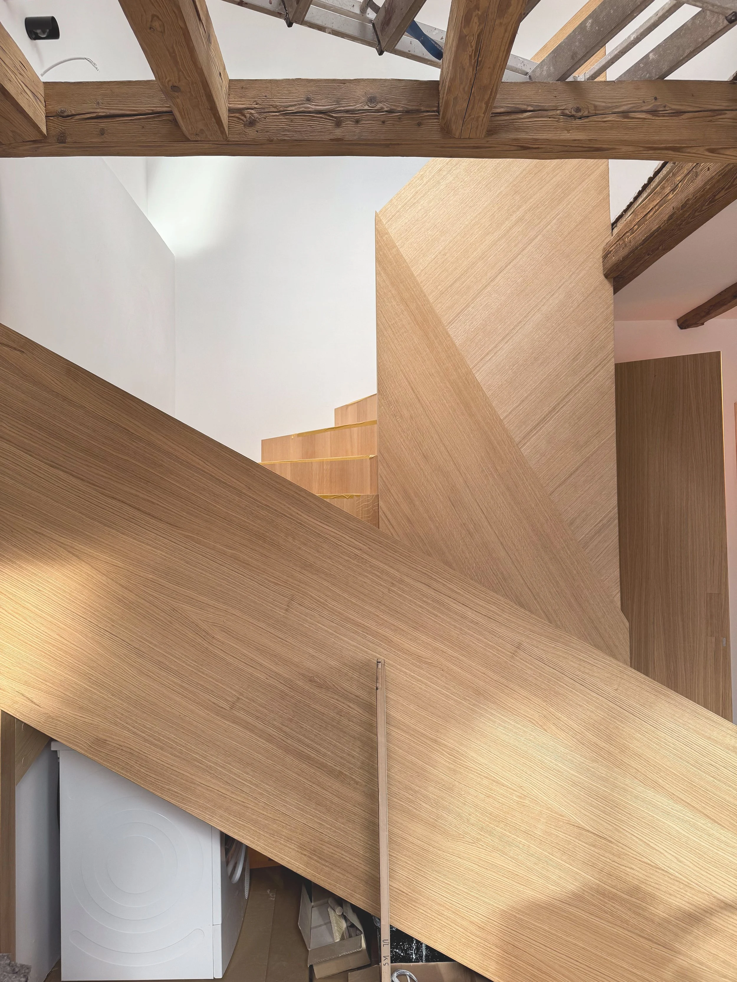 Moderne, skulpturale Holztreppe aus heller Eiche mit geschlossener Brüstung unter einer historischen Holzbalkendecke im Dachgeschoss.