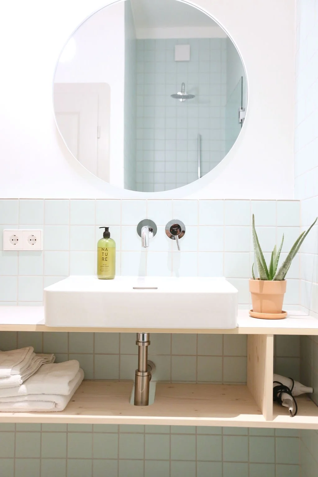 Modernes Badezimmer mit hellgrün gefliester Wand, Unterputz-Armatur, Aufsatzwaschbecken auf Holzwaschtisch und rundem Wandspiegel.