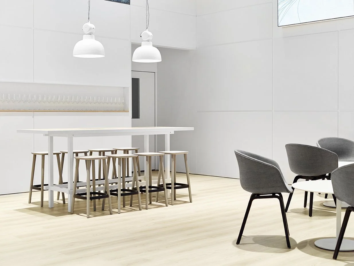 Heller Loungebereich auf dem Messestand mit hohem weißen Tisch, Holzhockern, zwei Hängeleuchten und grauen Polstersesseln an einem kleinen runden Tisch.