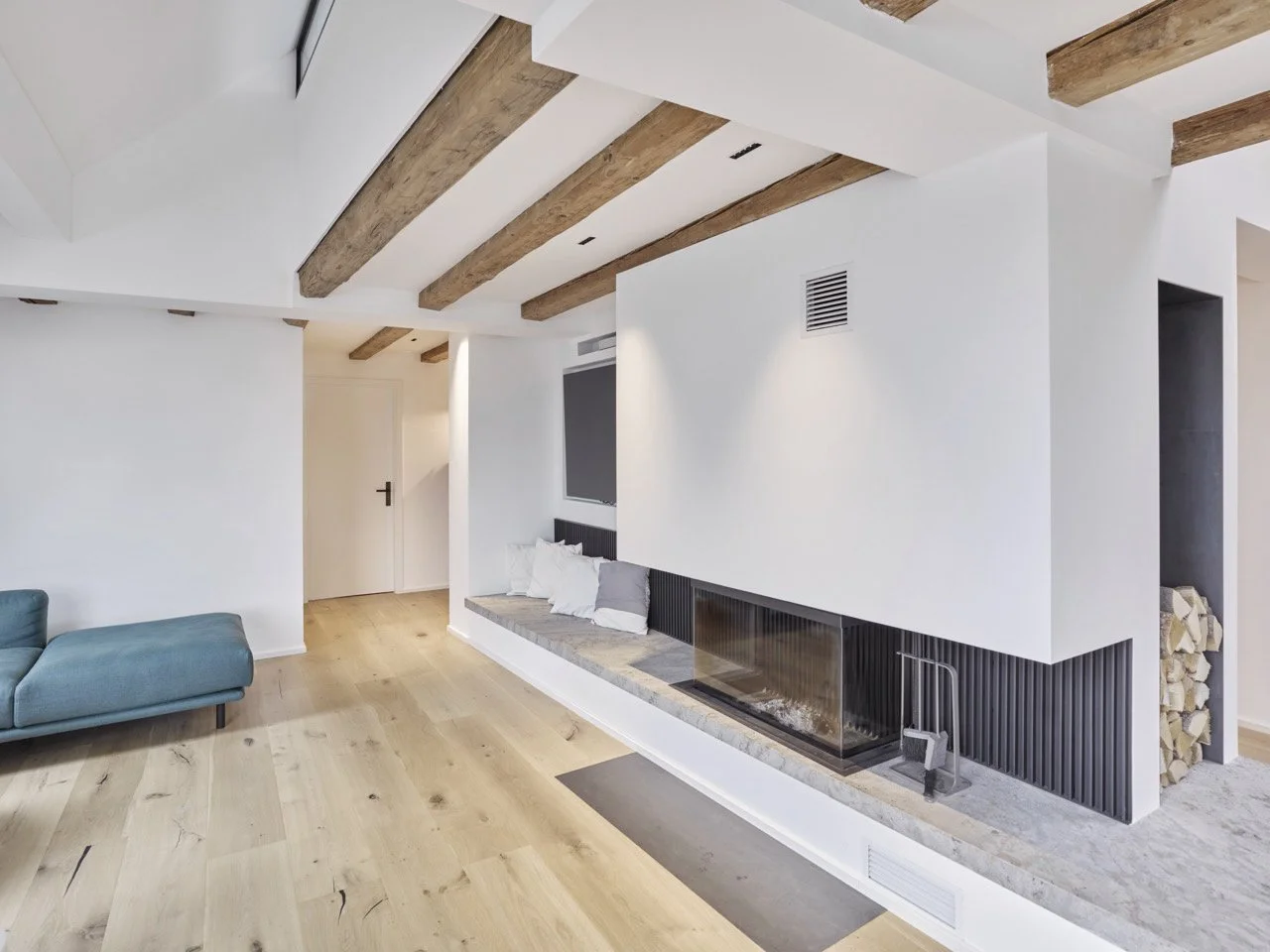 Offener Wohnbereich mit hellem Eichendielenboden, modernem Kamineinbau, integrierter Sitzbank, Holzlager-Nische und sichtbaren historischen Deckenbalken im Dachgeschoss-Projekt K7.