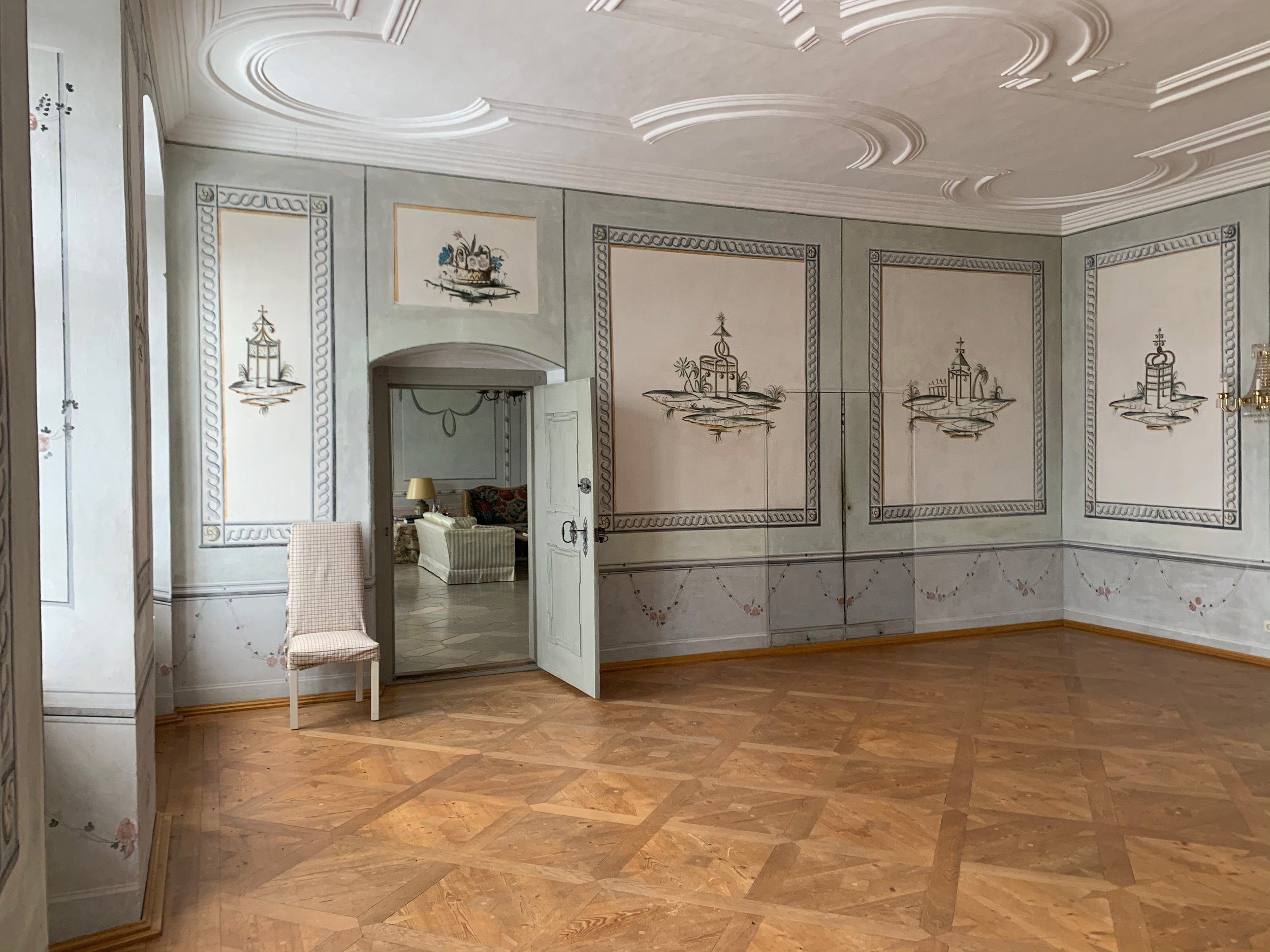 Restaurierter Prunksaal im Schloss mit historischen Wandmalereien, aufwendiger Rahmung, Barock-Stuckdecke, edlem Tafelparkett und Blick durch die geöffnete Flügeltür.