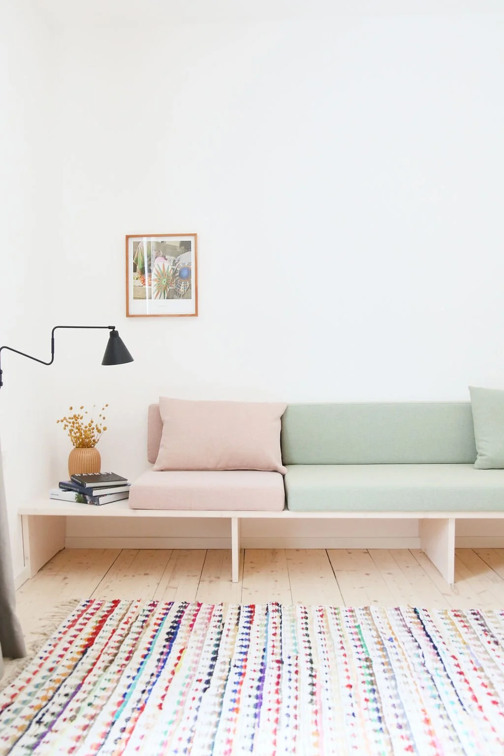 Minimalistischer Sitzbereich mit individuell gefertigter Holzbank mit pastellrosa und pastellgrünen Polstern, Dielenboden und bunter Webe-Laufmatte.