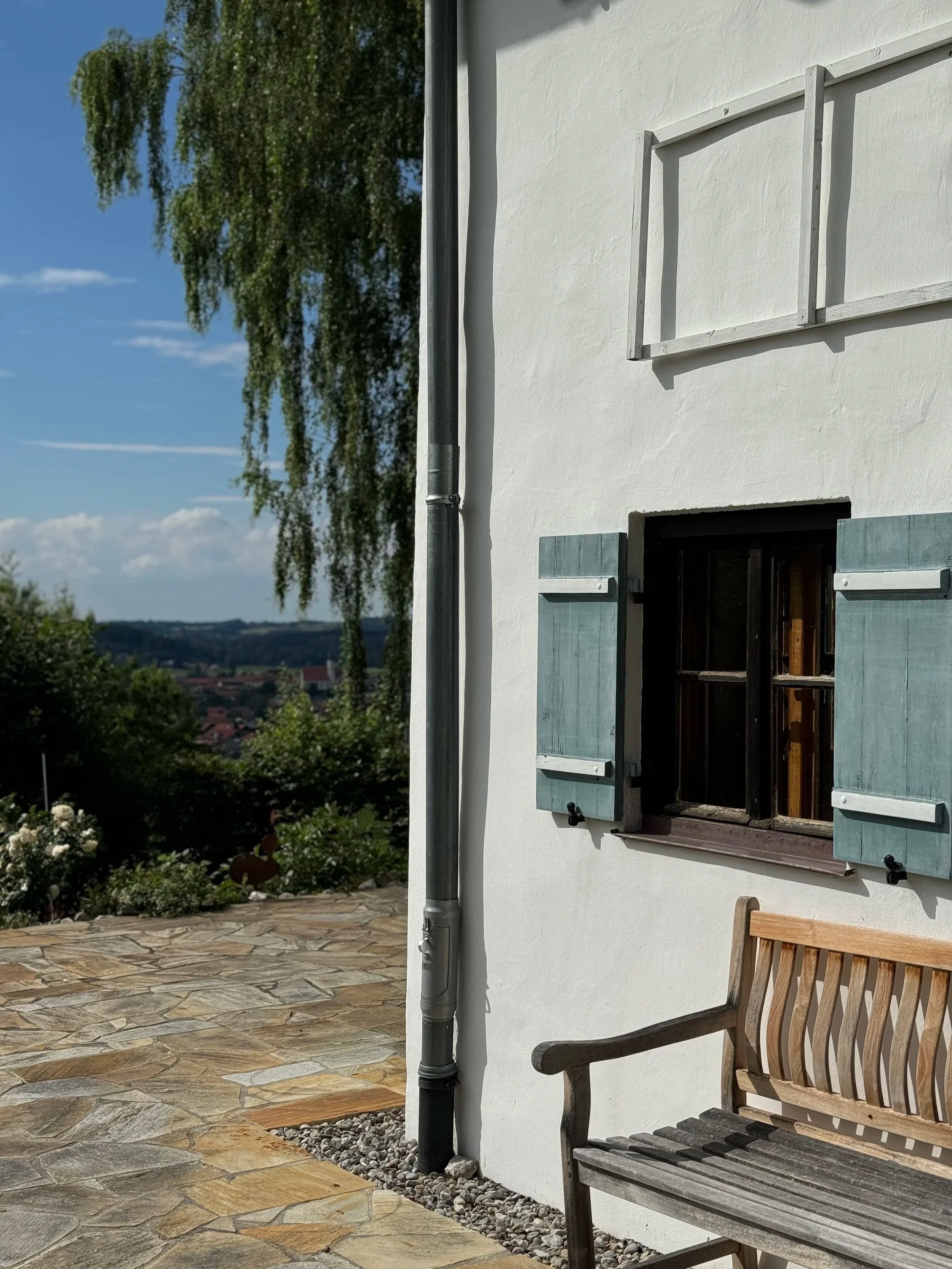 Detailaufnahme der Bestandsfassade eines alpenländischen Haupthauses mit hellblauen Holz-Klappläden, weißem Putz, einer Holzbank auf Natursteinpflaster und Weitblick.