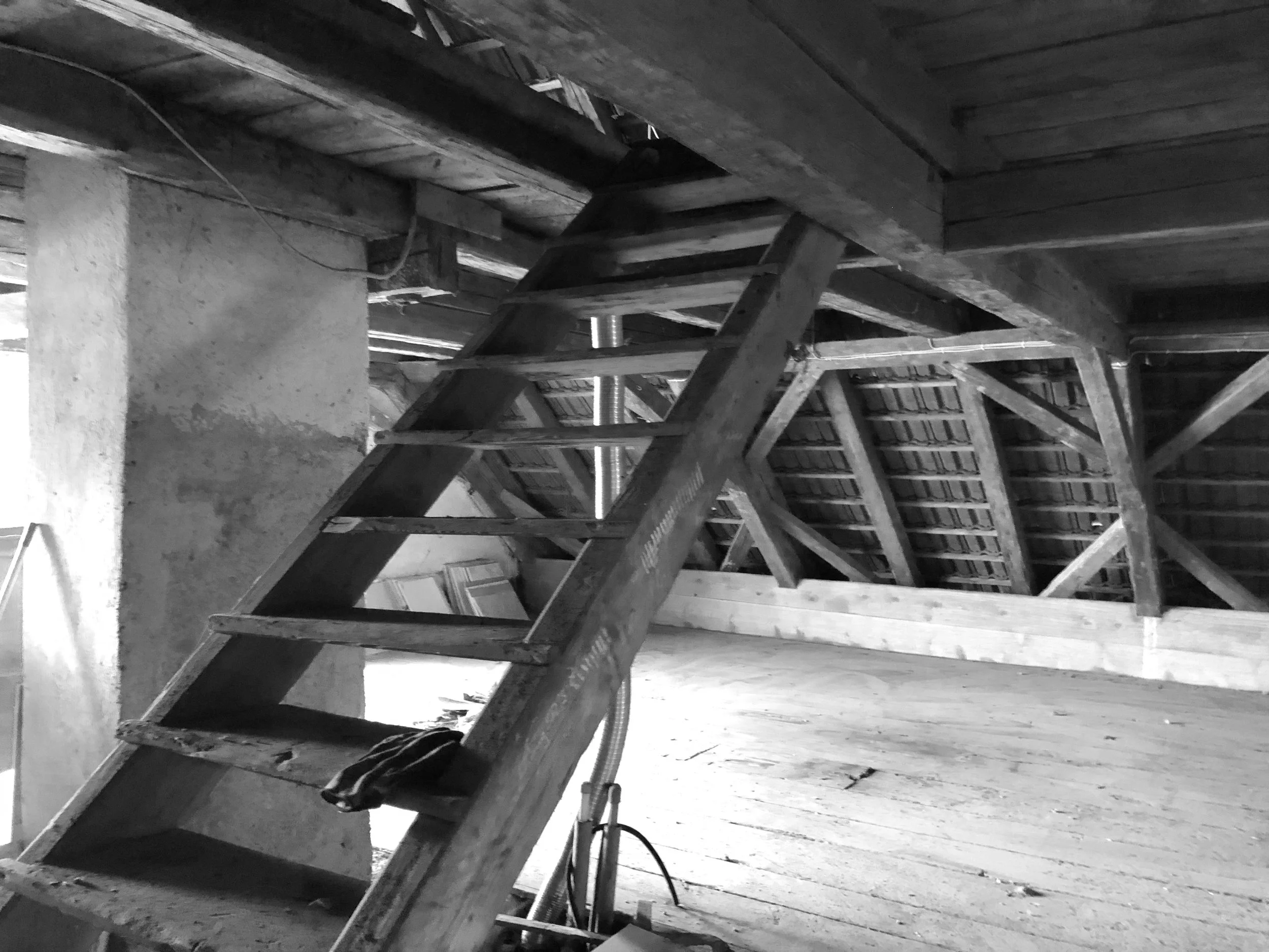 Schwarz-Weiß-Aufnahme des unrenovierten, alten Dachbodens vor dem Umbau mit rustikaler Holzstiege, offenem Dachstuhl und unfertigem Holzboden.