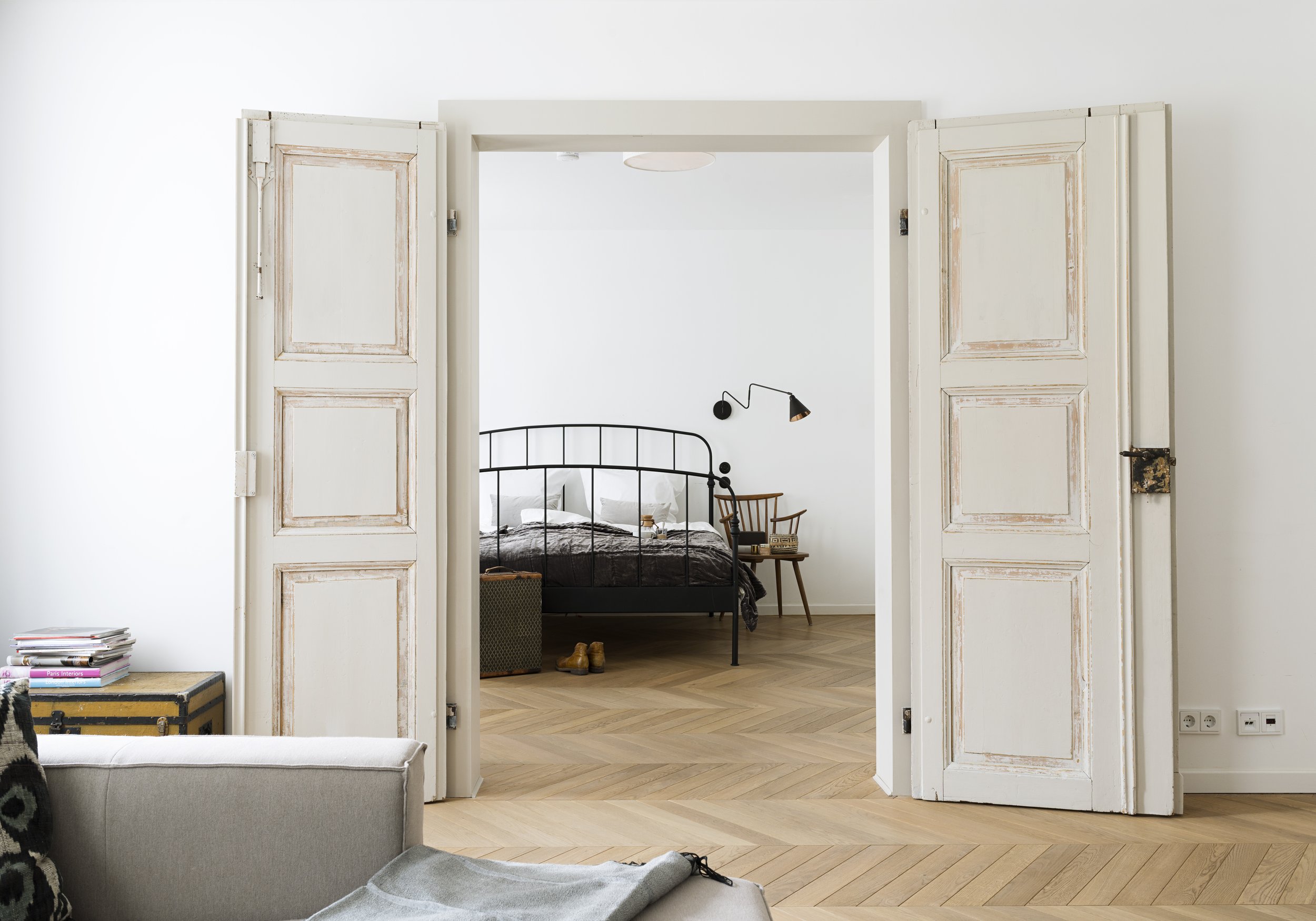 Blick durch eine offene, antike weiße Flügeltür in ein helles Schlafzimmer. Zu sehen ist ein schwarzes Metallbett, ein Holzstuhl mit Nachttisch, eine Wandleuchte und der durchgehende Fischgrät-Parkettboden.