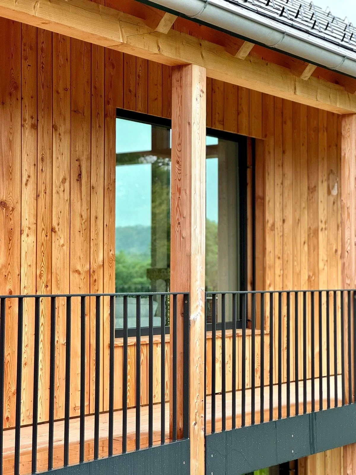 Nahaufnahme einer modernen Holzfassade mit vertikaler Lärcheschalung, Stützen, schwarzem Metallgeländer im Laubengang.