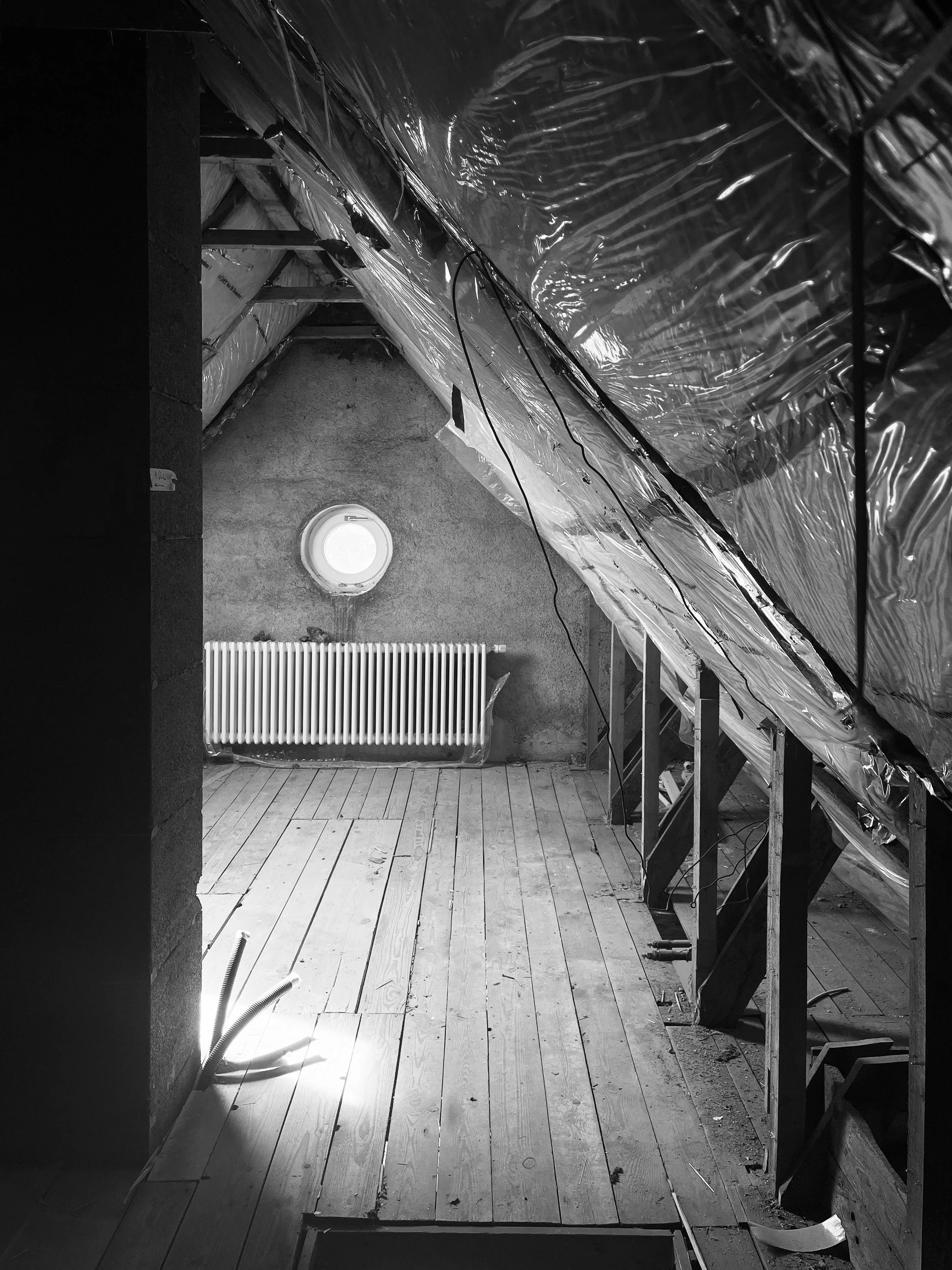 Aufnahme eines Dachgeschosses im Rohbau mit abgedichteter Folien-Dampfsperre an den Schrägen, altem Dielenboden, einem Heizkörper und einem kleinen Rundfenster (Ochsenauge) an der Stirnwand.