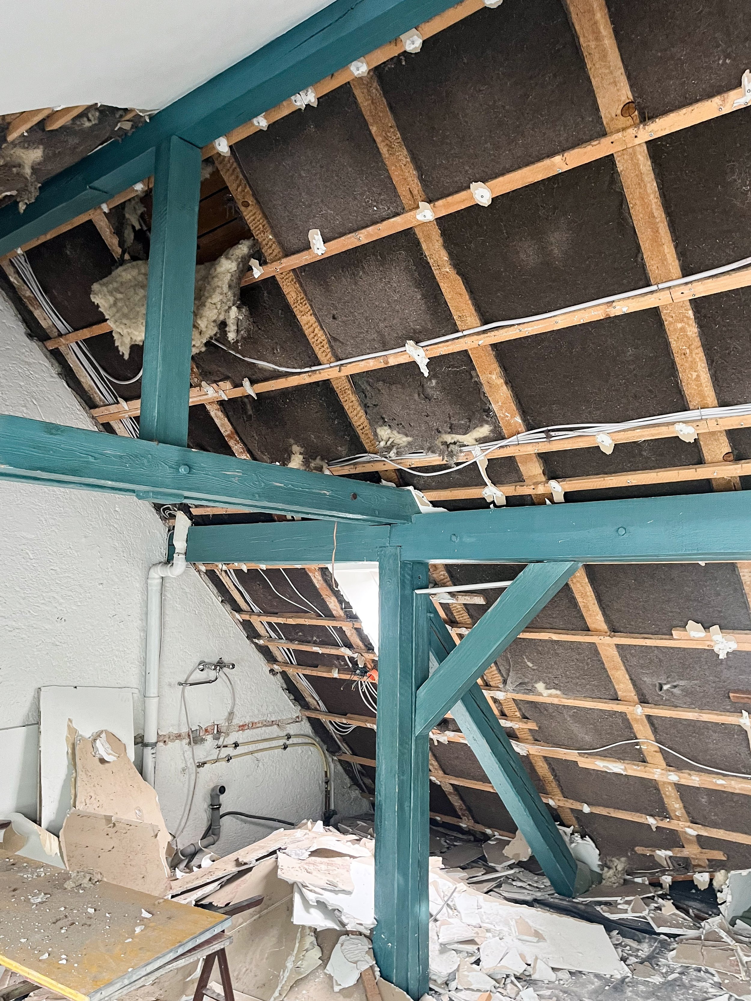 Eine Nahaufnahme einer Baustelle im Dachgeschoss. Die Decken- und Dachkonstruktion ist teilweise freigelegt und zeigt die Holzbalken (Türkisblau gestrichen), Dämmmaterial und die Holzlattung.