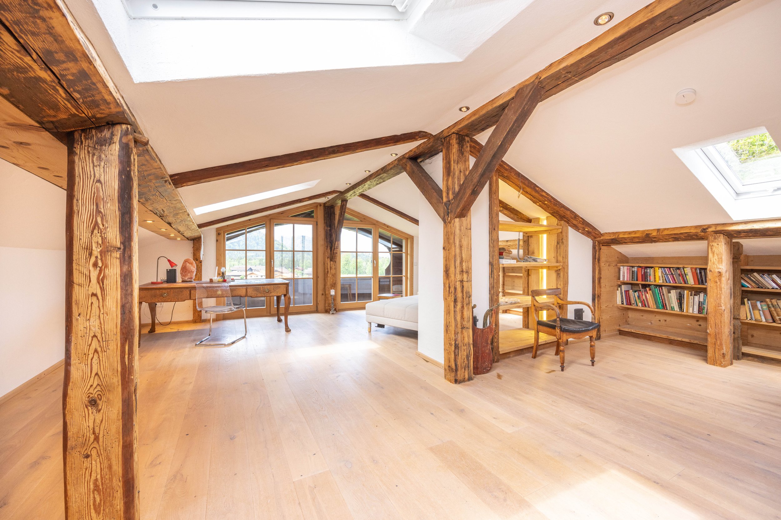 Ausgebautes Dachgeschoss mit freigelegtem Holzgebälk, hellmöglichem Parkettboden, Velux-Dachfenstern, eingebauten Bücherregalen und Schreibtisch vor der Verglasung.