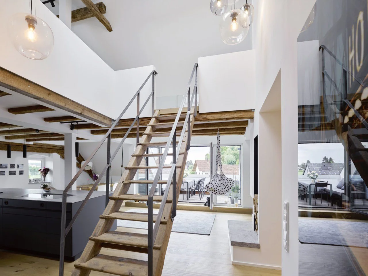 Offenes, zweigeschossiges Dachgeschoss mit rustikaler Holztreppe, dunkler Kücheninsel, sichtbaren Holzbalken und Blick durch die Glasfront auf die Dachterrasse im Projekt K7 in Fürstenfeldbruck.