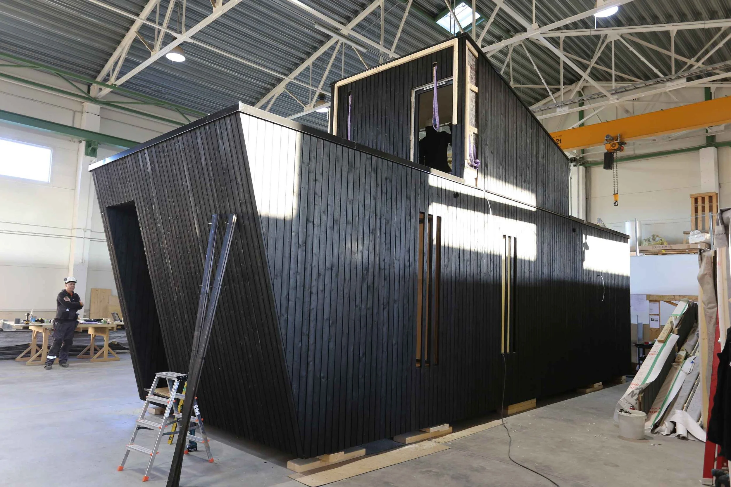 Werkstattfertigung eines modernen Modulhauses mit vertikaler, dunkler Holzschalung und schrägem Wandelement in einer Industriehalle mit Bauarbeiter im Hintergrund.