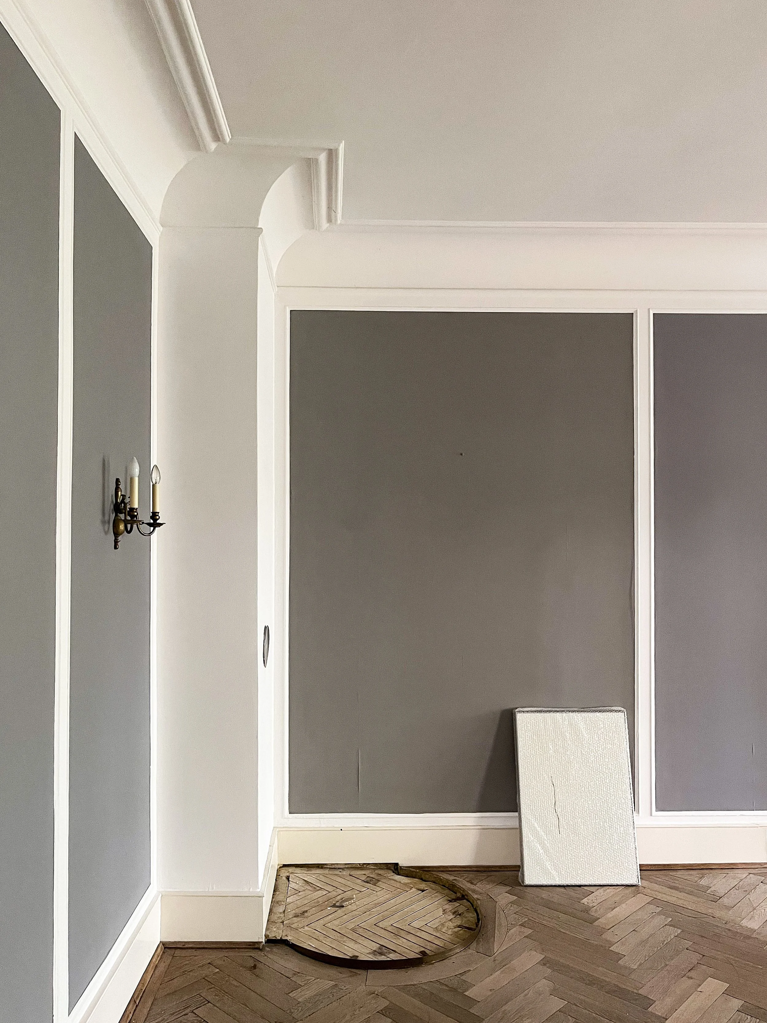 Innenraum-Ecke mit Fischgrät-Parkett, grauen Wänden und weißer Wandtäfelung. Eine antike Messing-Leuchte und eine Heizkörperverkleidung sind zu sehen.