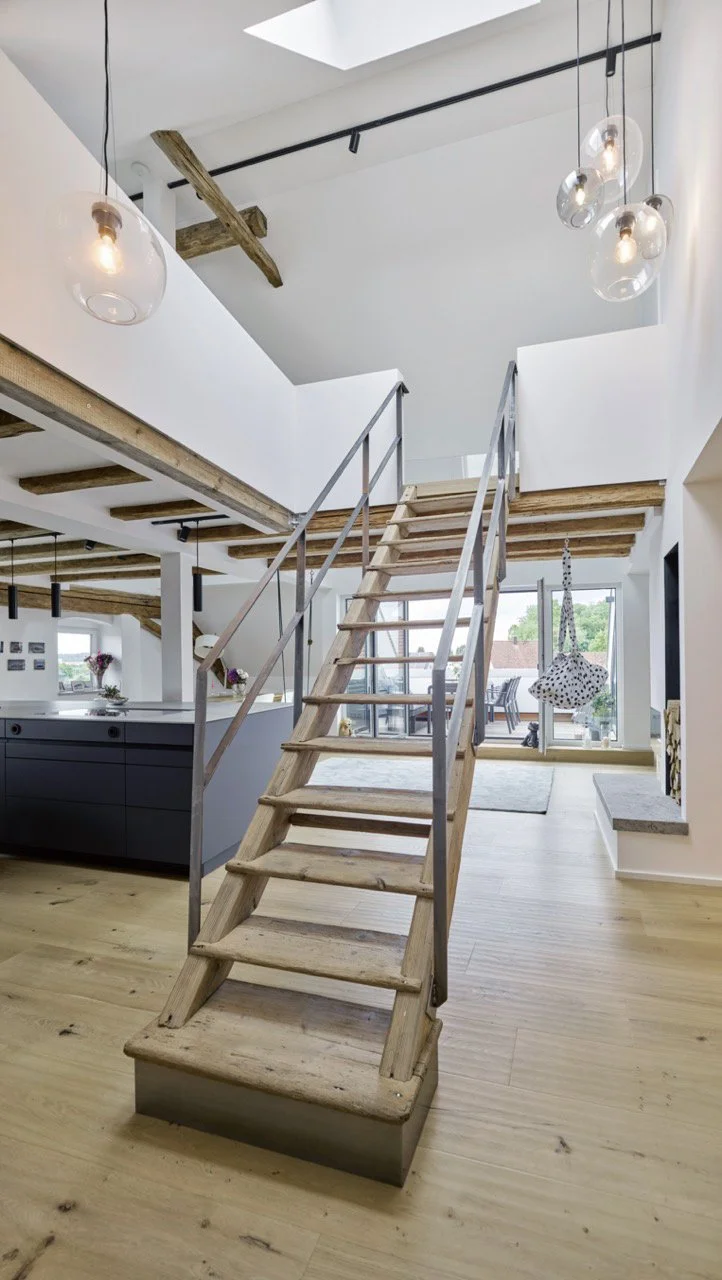 Frontale Ansicht einer maßgefertigten Holztreppe mit Metallgeländer, die im ausgebauten Dachgeschoss zur Empore führt, umgeben von Holzdielenboden, Glaskugelleuchten und sichtbaren Deckenbalken.
