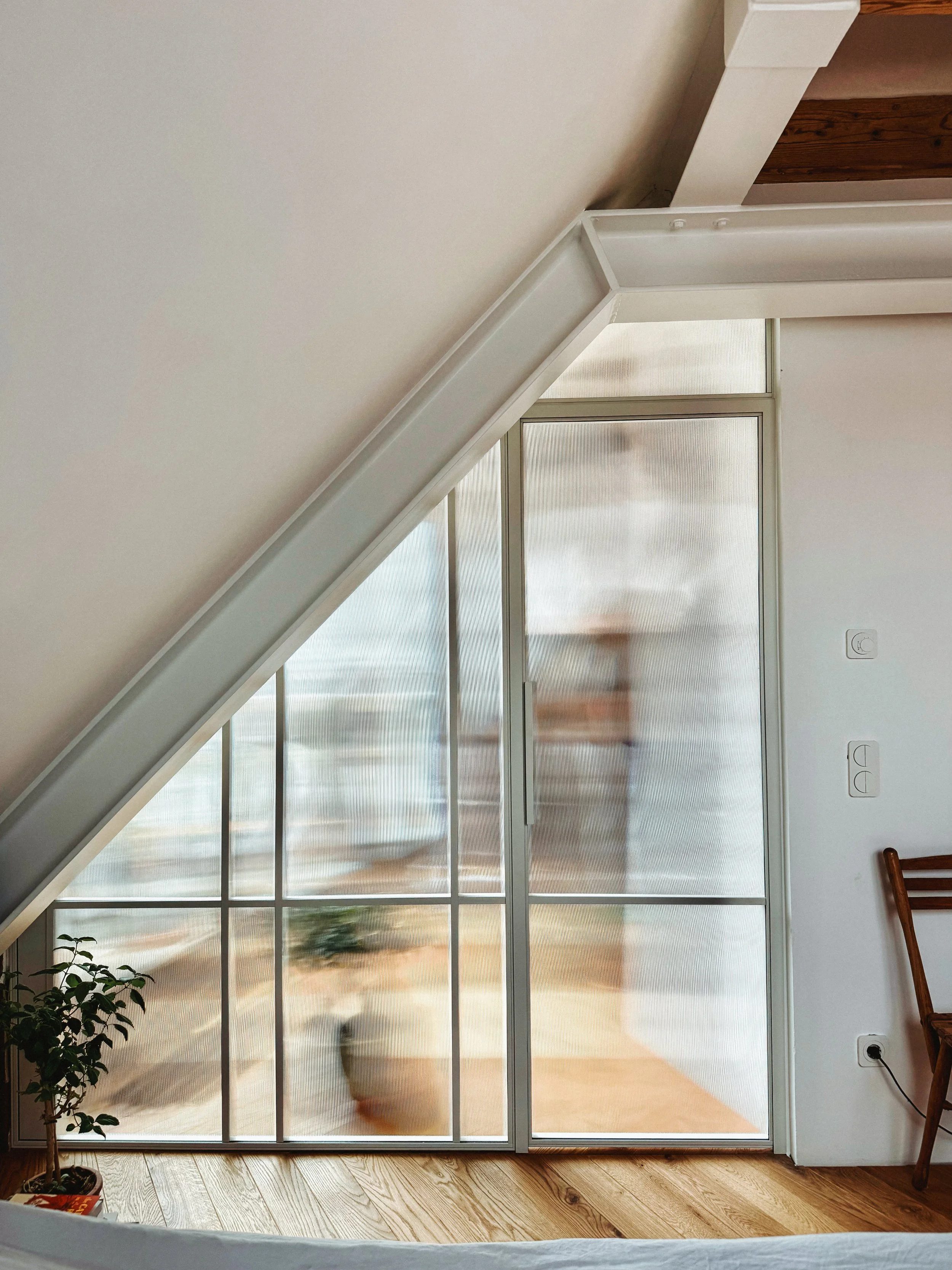 Maßgefertigte Glas-Stahl-Trennwand mit Riffelglas und Schiebetür, präzise eingepasst in eine Dachschräge unter weiß lackierten Stahlträgern im modernen Dachausbau.