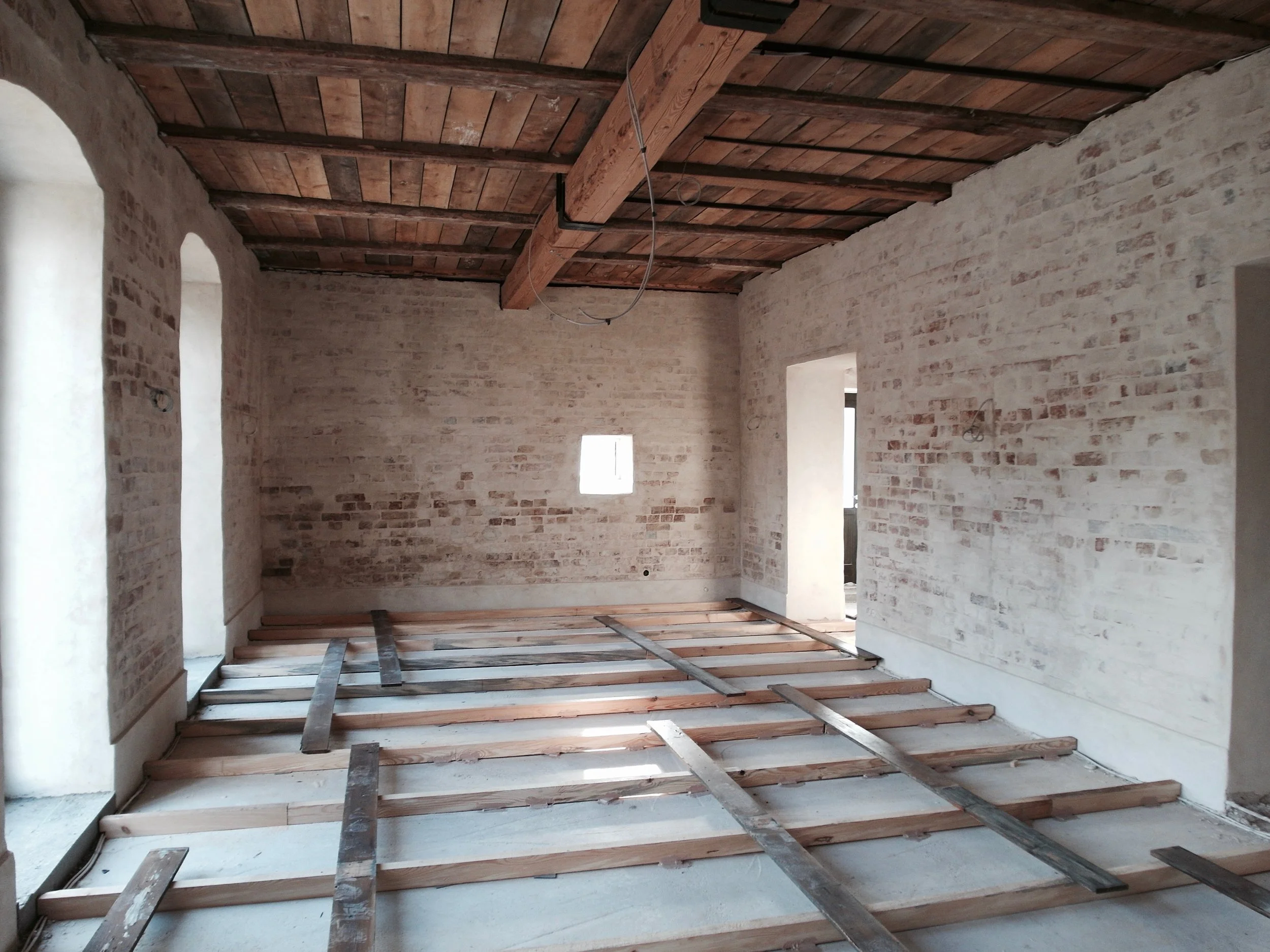 Perspektivische Ansicht eines großen, rohen Raumes in einem historischen Gebäude. Zu sehen sind eine massive Holzbalkendecke, weiß geschlämmtes Backsteinmauerwerk und eine Holzunterkonstruktion für den neuen Dielenboden.