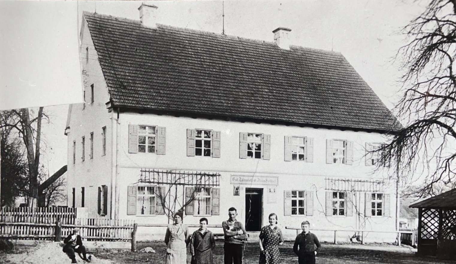Historisches Schwarz-Weiß-Foto des mehr als 100 Jahre alten Bestandsgebäudes mit Satteldach, Klappläden an den Fenstern und einer Gruppe von Menschen vor dem Haus.