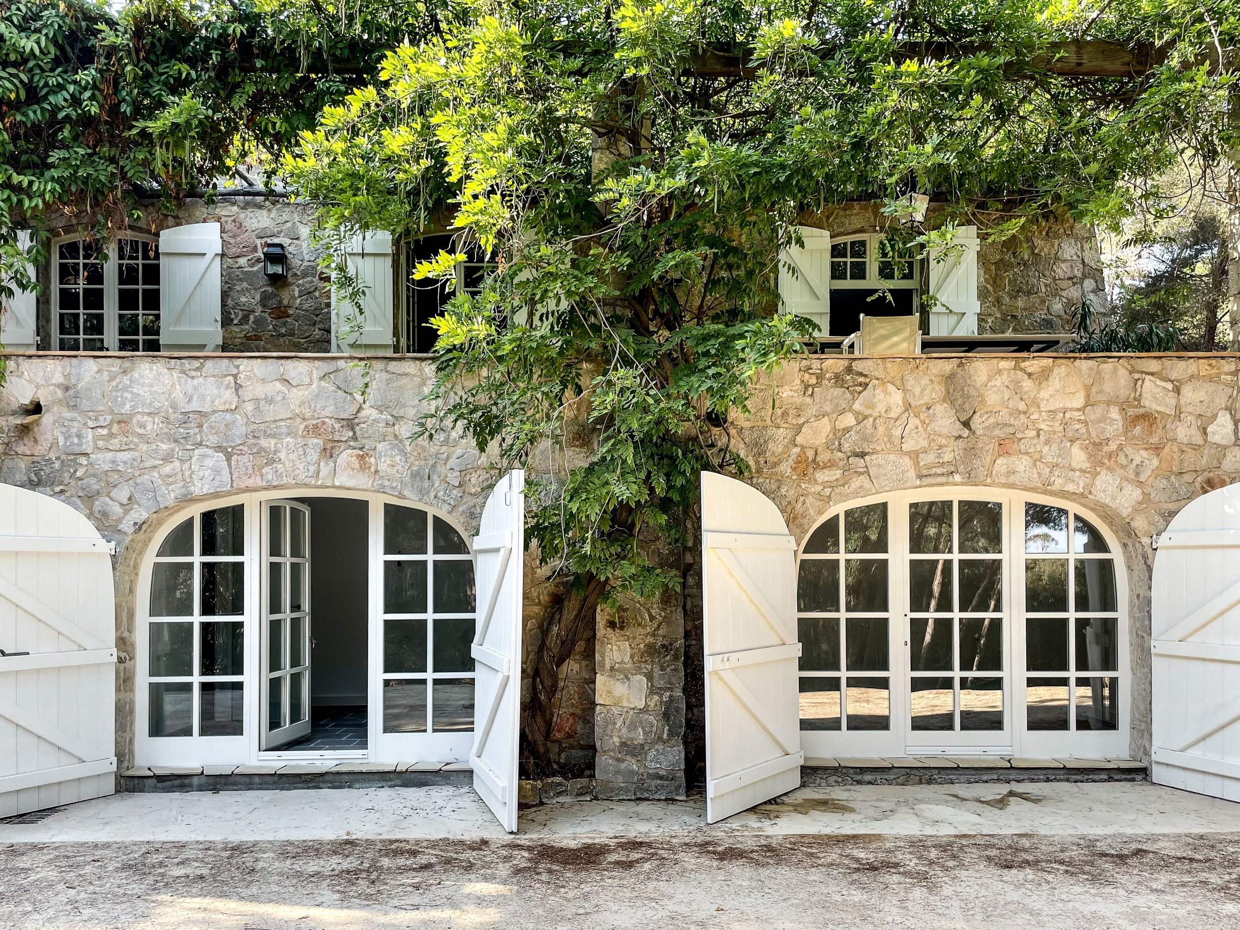 Rustikale Naturstein-Fassade eines Sommerhauses in St. Tropez mit zwei großzügigen Rundbogen-Sprossenfenstern und geöffneten, weiß lackierten Holz-Klappläden.