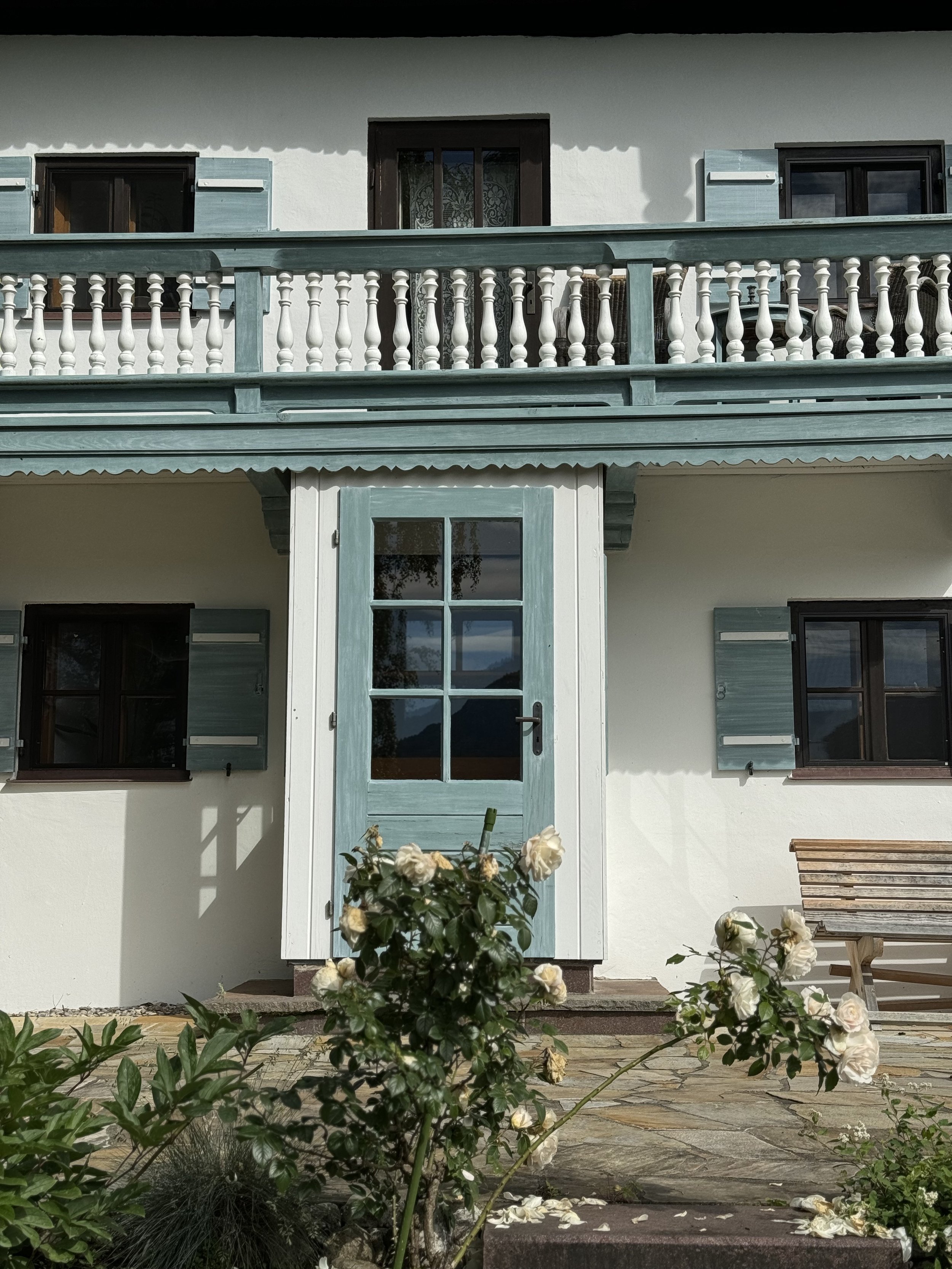 Frontalansicht der Haupthaus-Bestandsfassade mit hellblauem Söller (Balkon) und passender hellblauer Eingangstür mit Sprossenverglasung, flankiert von weißen Rosensträuchern.