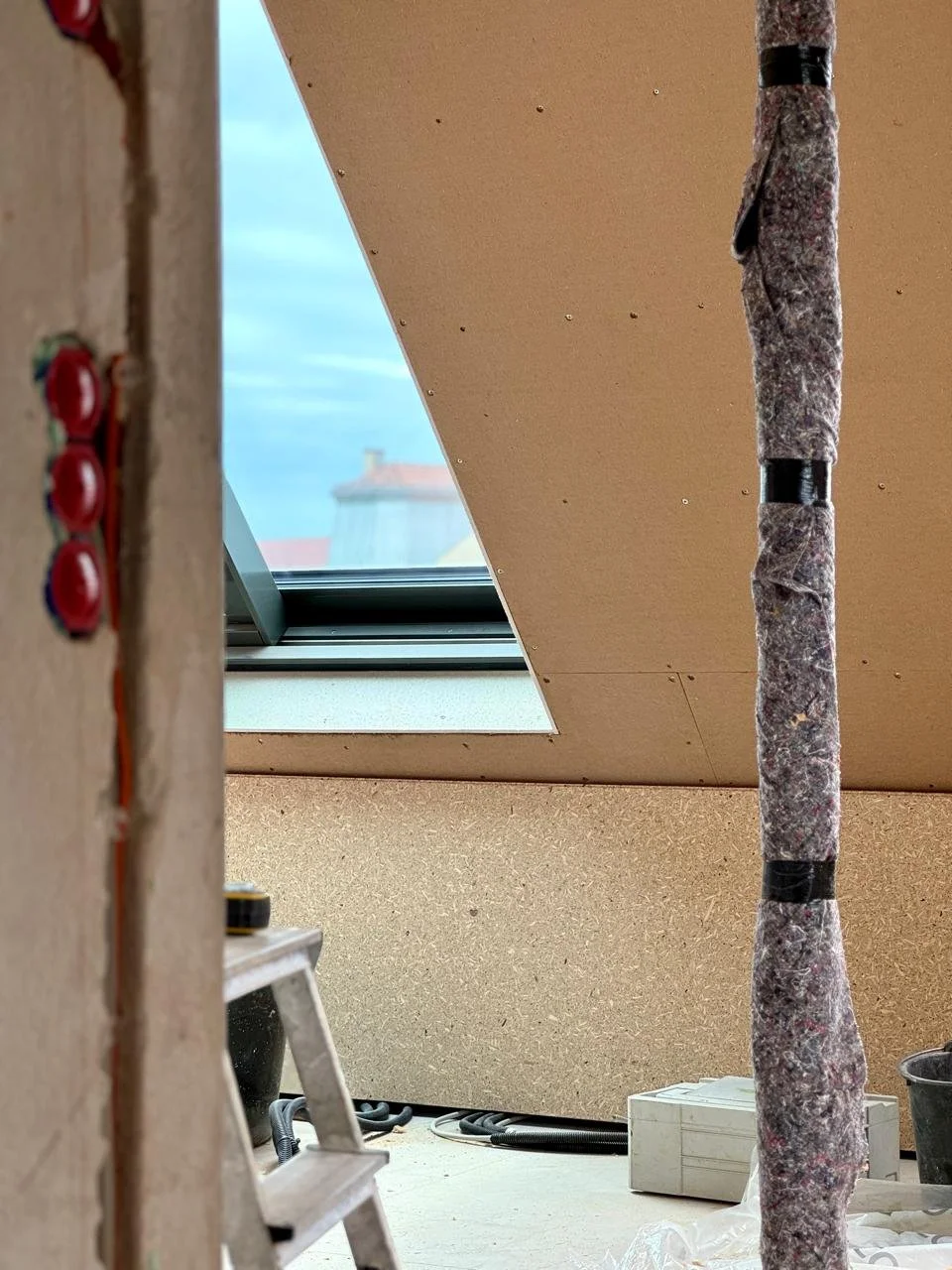 Detailaufnahme einer mit Dämmfilz umwickelten Fallleitung im Dachgeschoss einer Maisonette-Wohnung in München neben einer Wandschräge mit Blick aus dem Dachfenster.