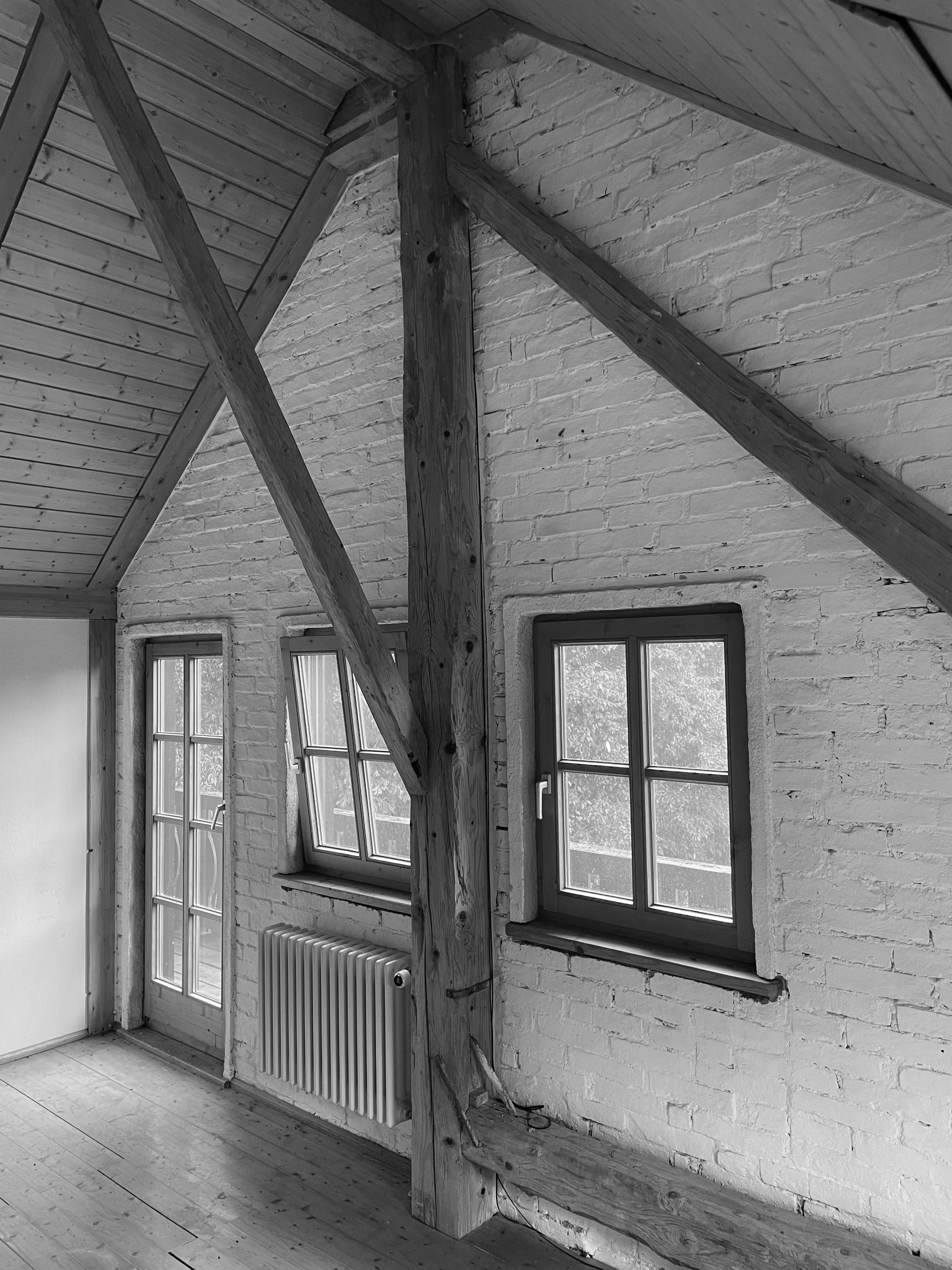 Aufnahme eines ausgebauten Dachgeschosses mit freigelegtem Holztragwerk, weiß gestrichener Backsteinwand, Holzsprossenfenstern und Dielenboden.