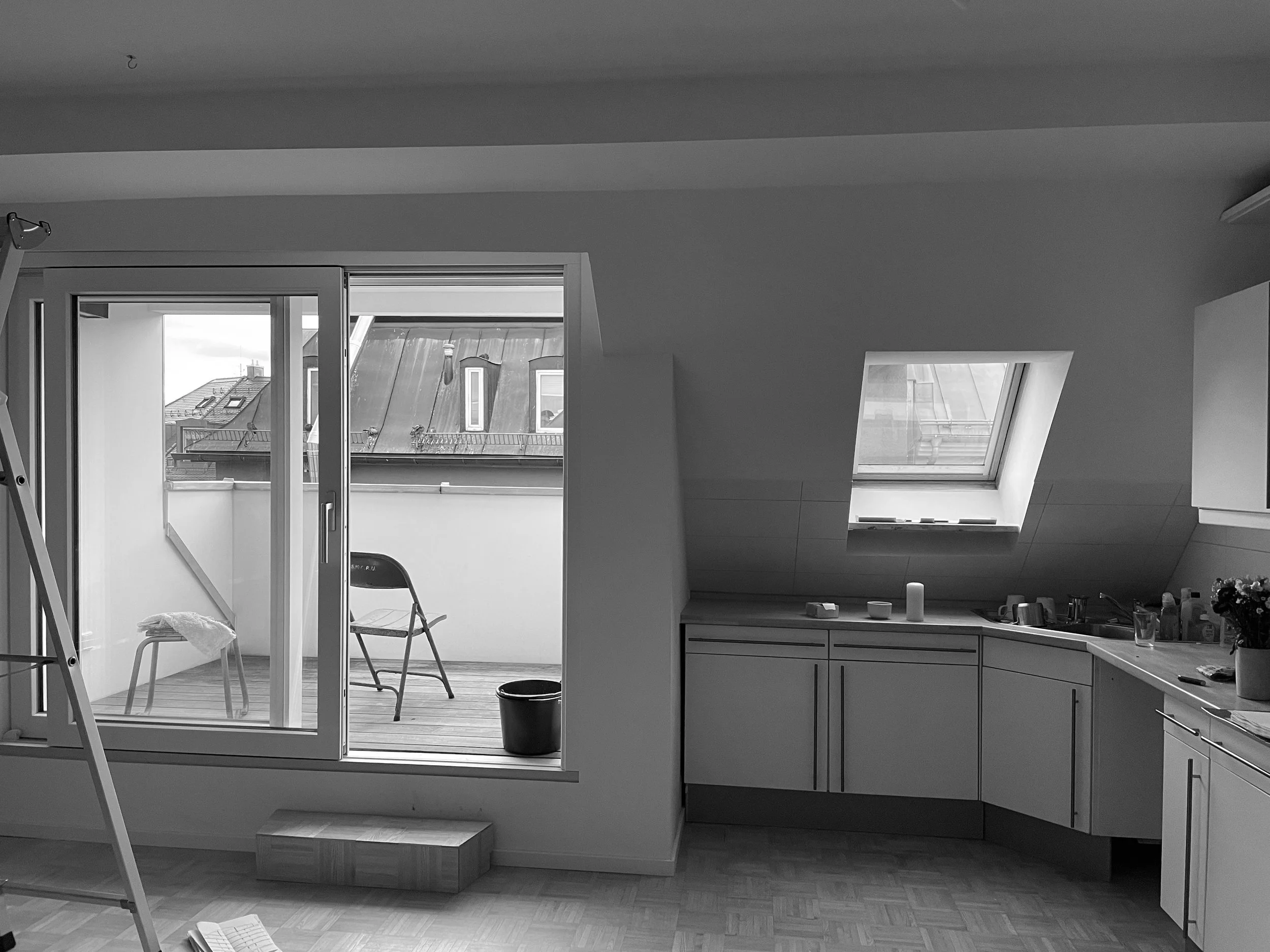 Dachgeschossraum vor der Sanierung mit Küche und Zugang zu einer kleinen Dachterrasse. Schräge Dachflächen mit Dachfenstern, Einbauküche und Parkettboden prägen den Raum.