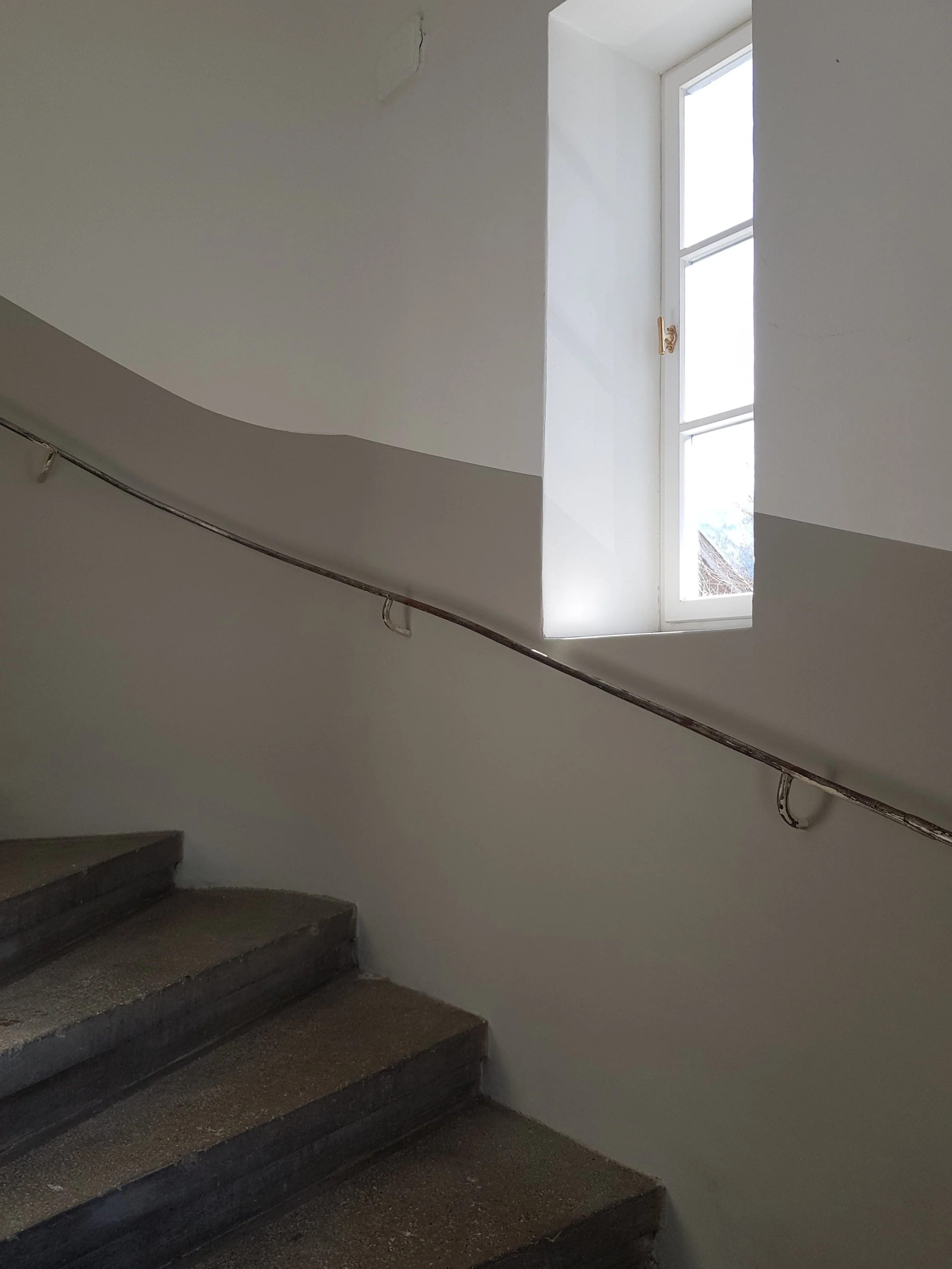 Nahaufnahme eines gewendelten Treppenlaufs im historischen Bestand mit sanft geschwungenem Wandanschnitt, langem Metallhandlauf und tief sitzendem, weißem Holz-Sprossenfenster.